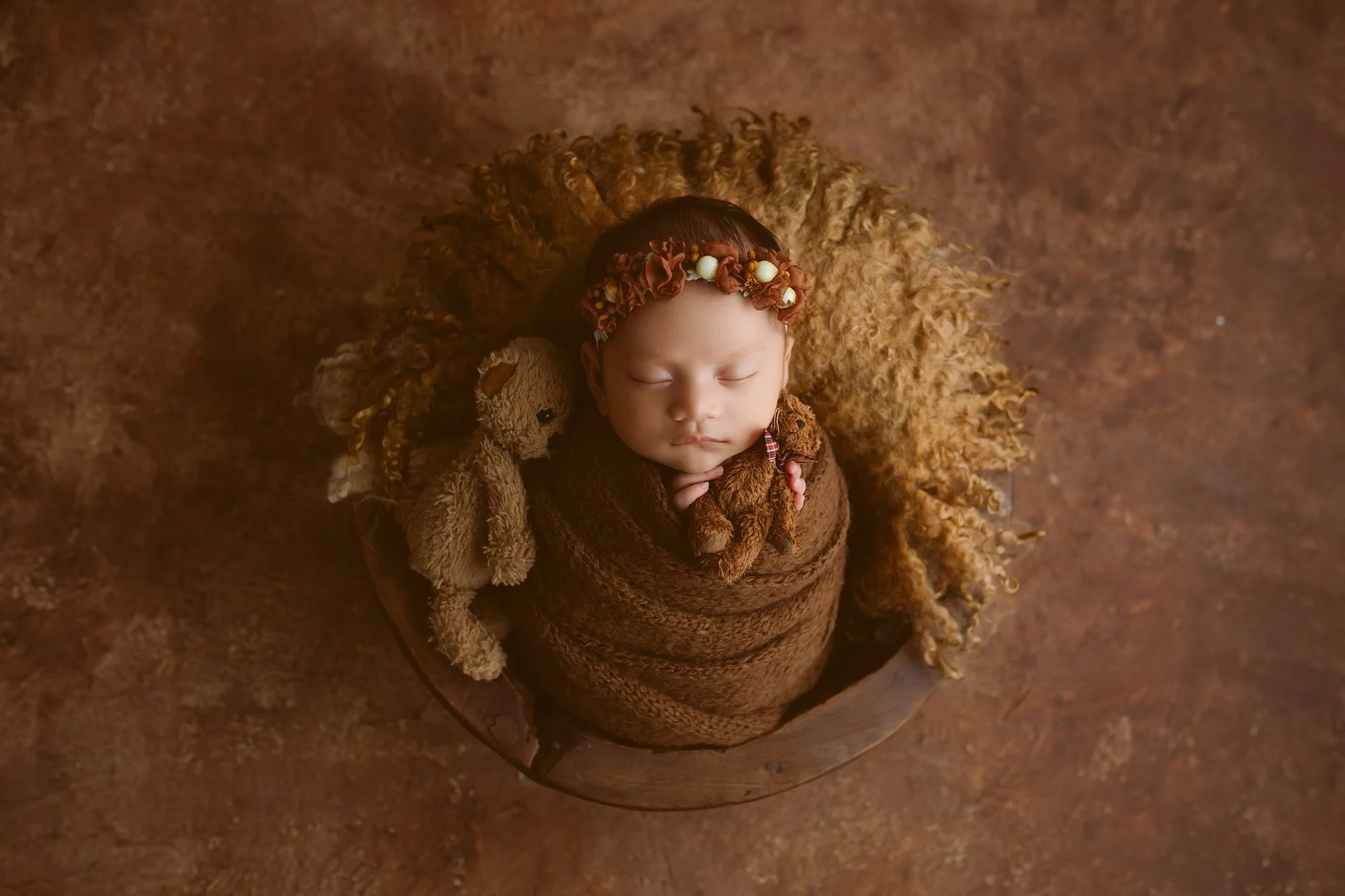 A sleeping baby wrapped in a brown blanket, wearing a floral headband, lying on a round wooden surface surrounded by plush teddy bears and a curly faux fur blanket, on a textured brown background.