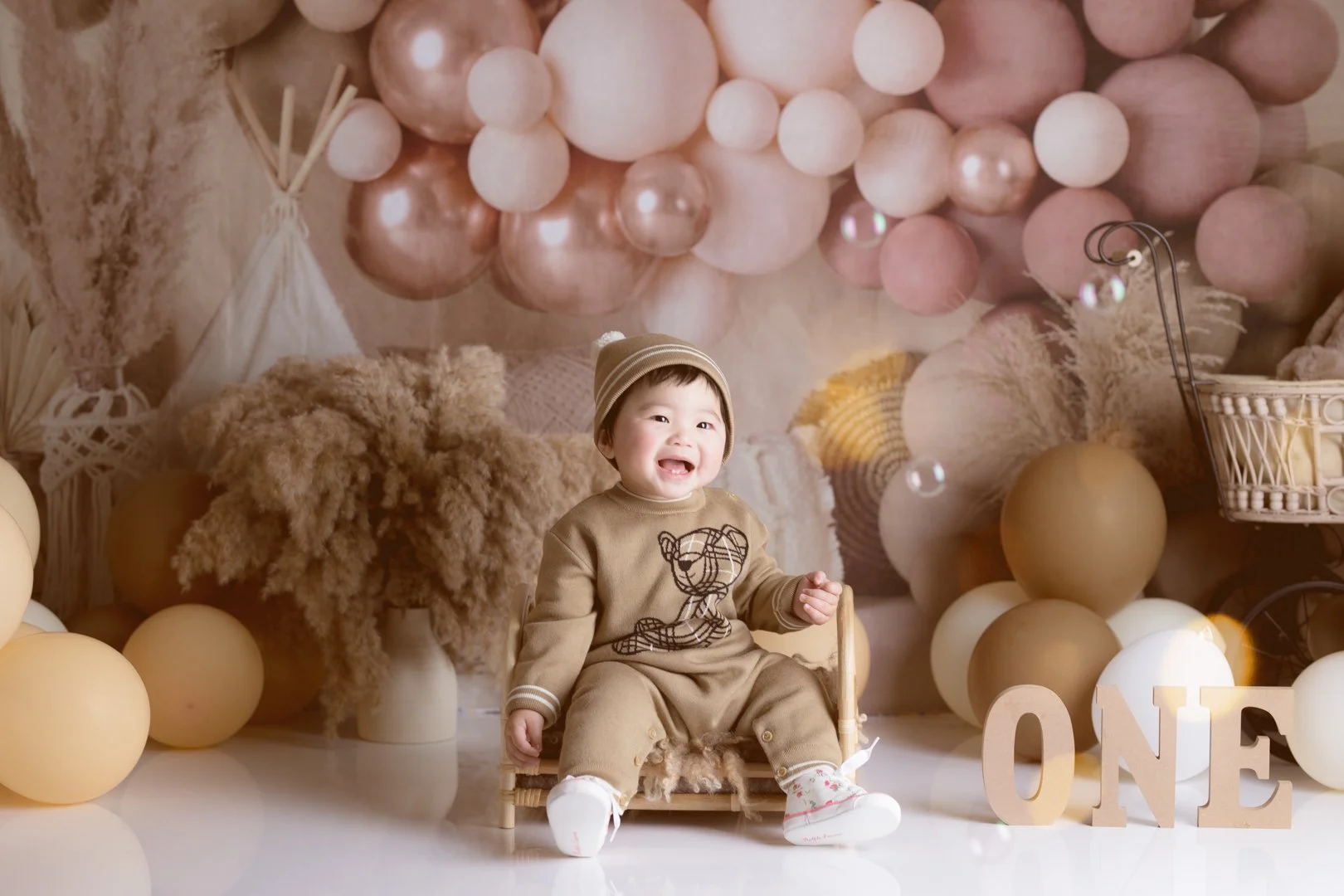 A cheerful  one year old girl sitting on a small chair, surrounded by beige and brown balloons, decorative dried plants, and a large 'ONE' sign, with a peach-colored balloon backdrop.
