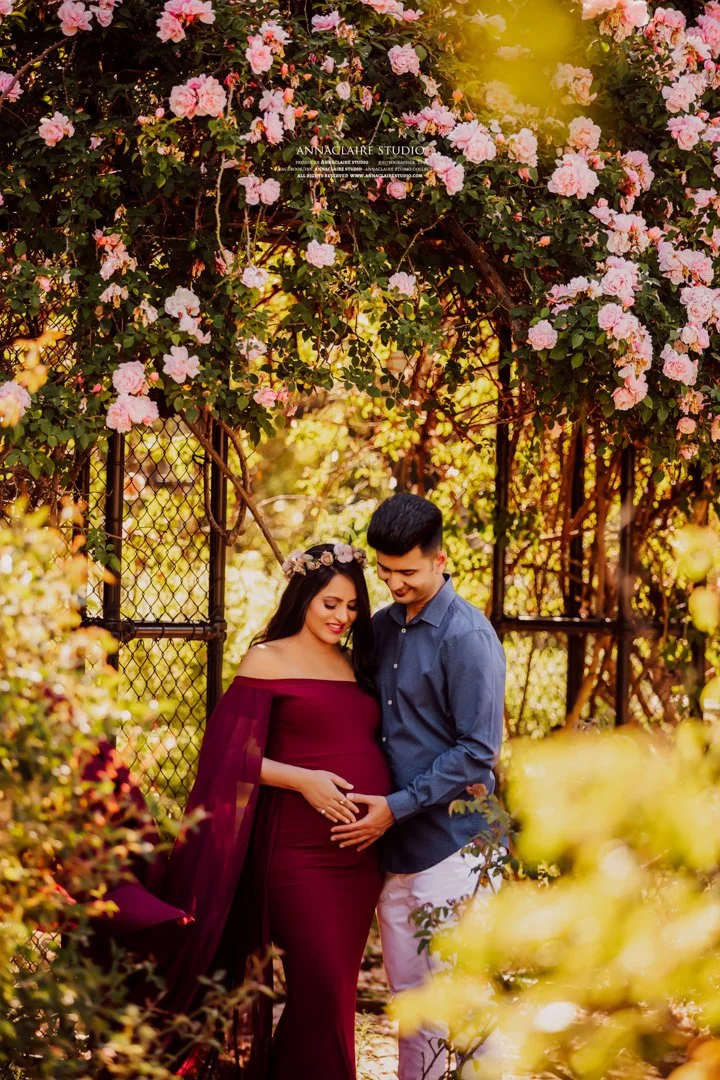 Pregnant woman in a burgundy off-shoulder dress stands with a man in a blue button-up shirt outside, surrounded by pink and yellow flowers, under a flowering arch.