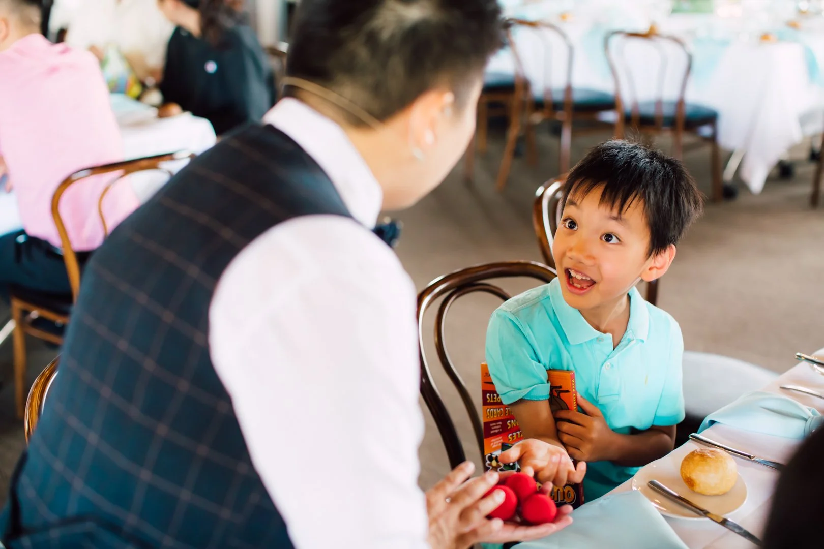 A young boy with Black hair and a light blue polo shirt smiles and talks to a waiter who is dressed formally in a white shirt and dark vest, holding a red plush toy and a colorful cocoa box, in a restaurant.