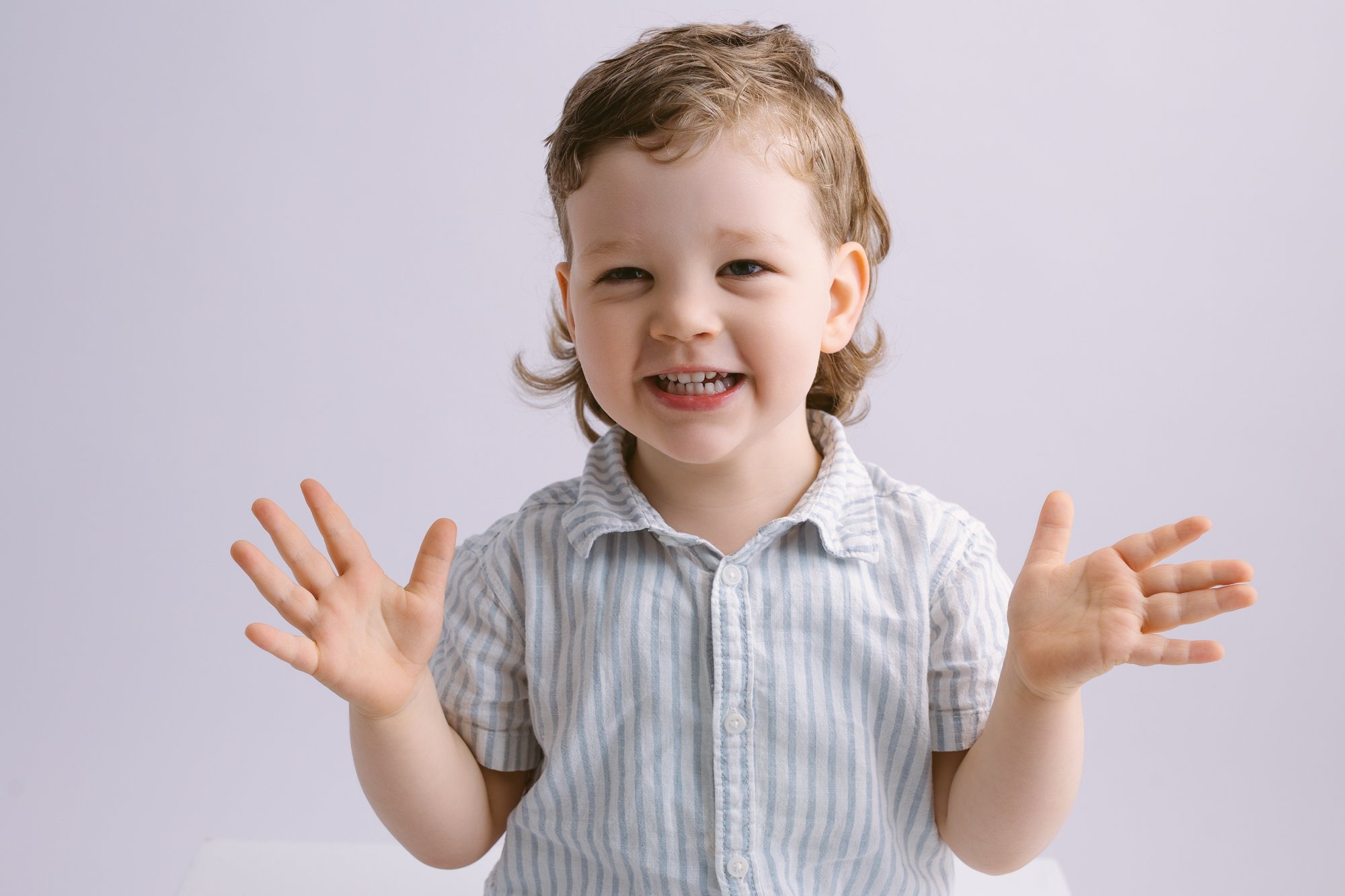 A young boy with brown curly hair smiling and playfully raising his hands, wearing a light-colored, striped button-up shirt against a plain white background.