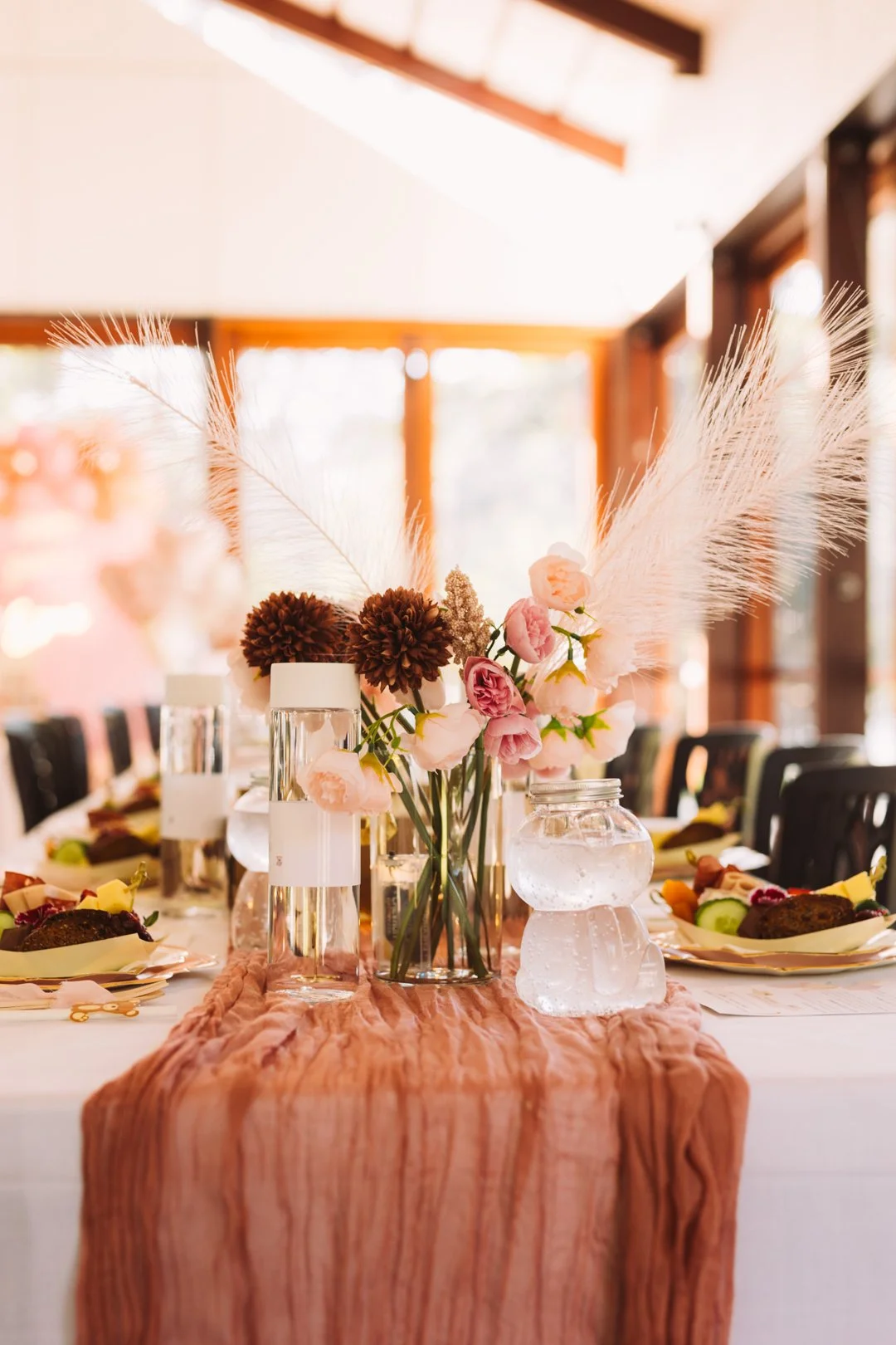 Elegant banquet table with a pink, textured table runner, floral centerpiece with pink and brown flowers and white feathers, surrounded by place settings with plates and food, in a warmly lit room with large windows.