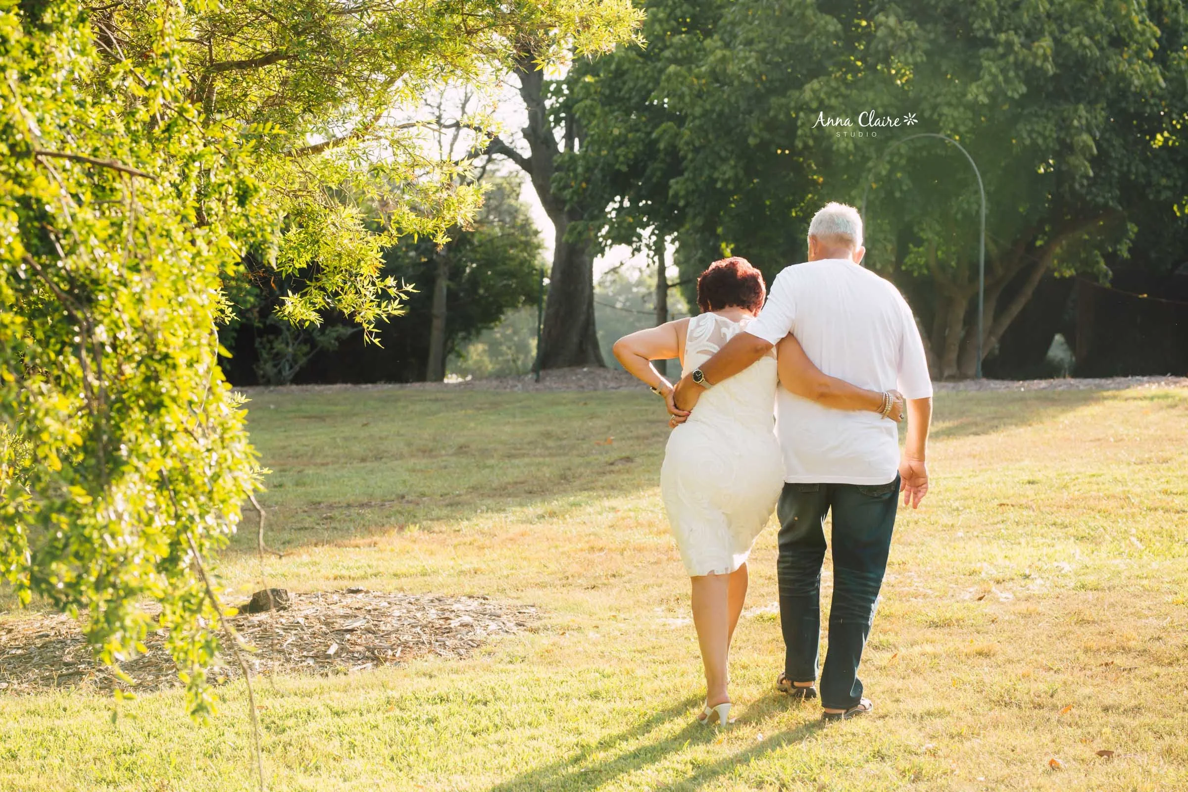 A couple walking together on a sunny grassy park, with trees in the background, embracing each other from behind, during late afternoon or early evening.