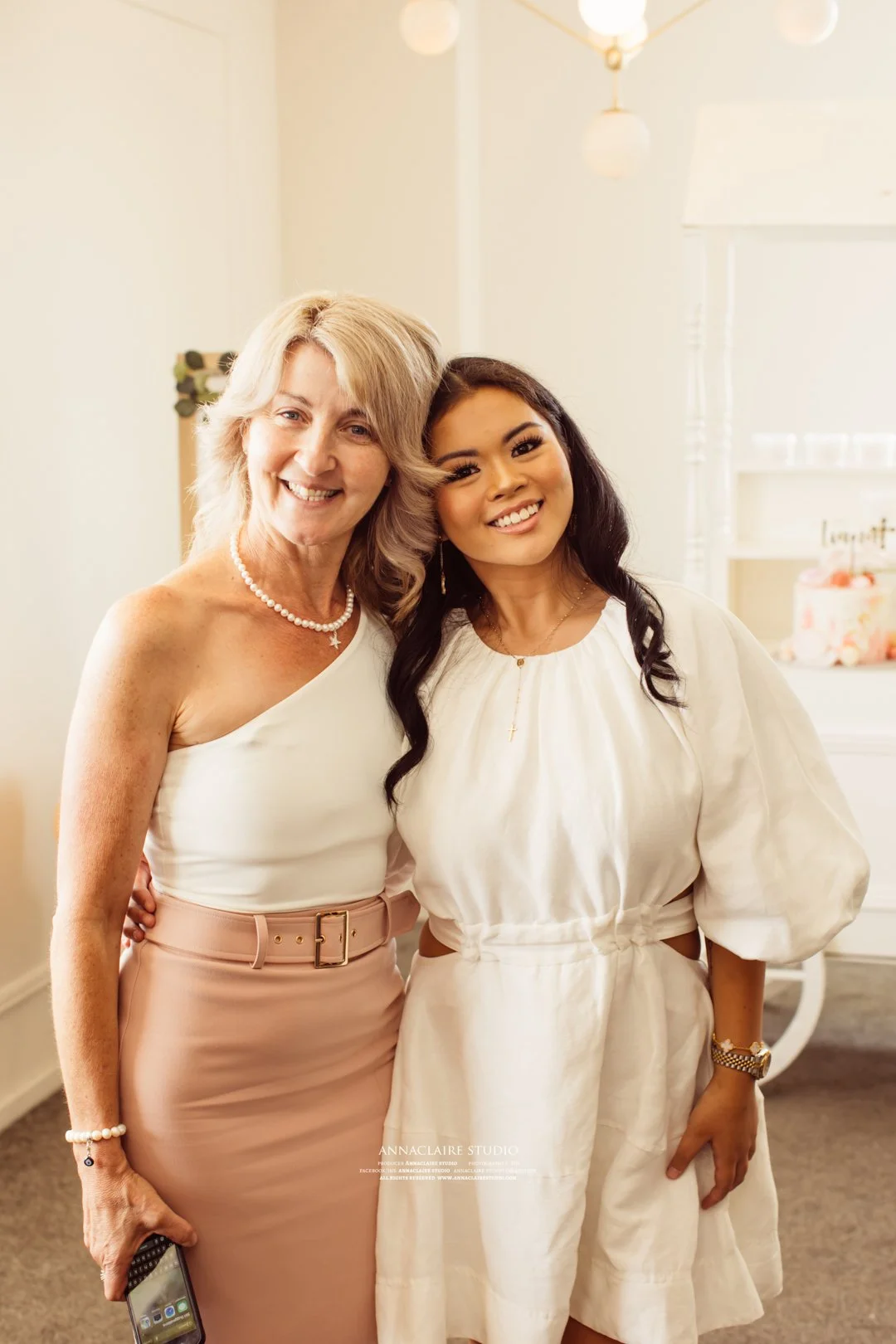 Two women standing close together indoors, smiling at the camera. One woman has blonde hair and is wearing a white one-shoulder top with a peach-colored skirt, holding a phone in her right hand. The other woman has dark hair and is wearing a cream-co