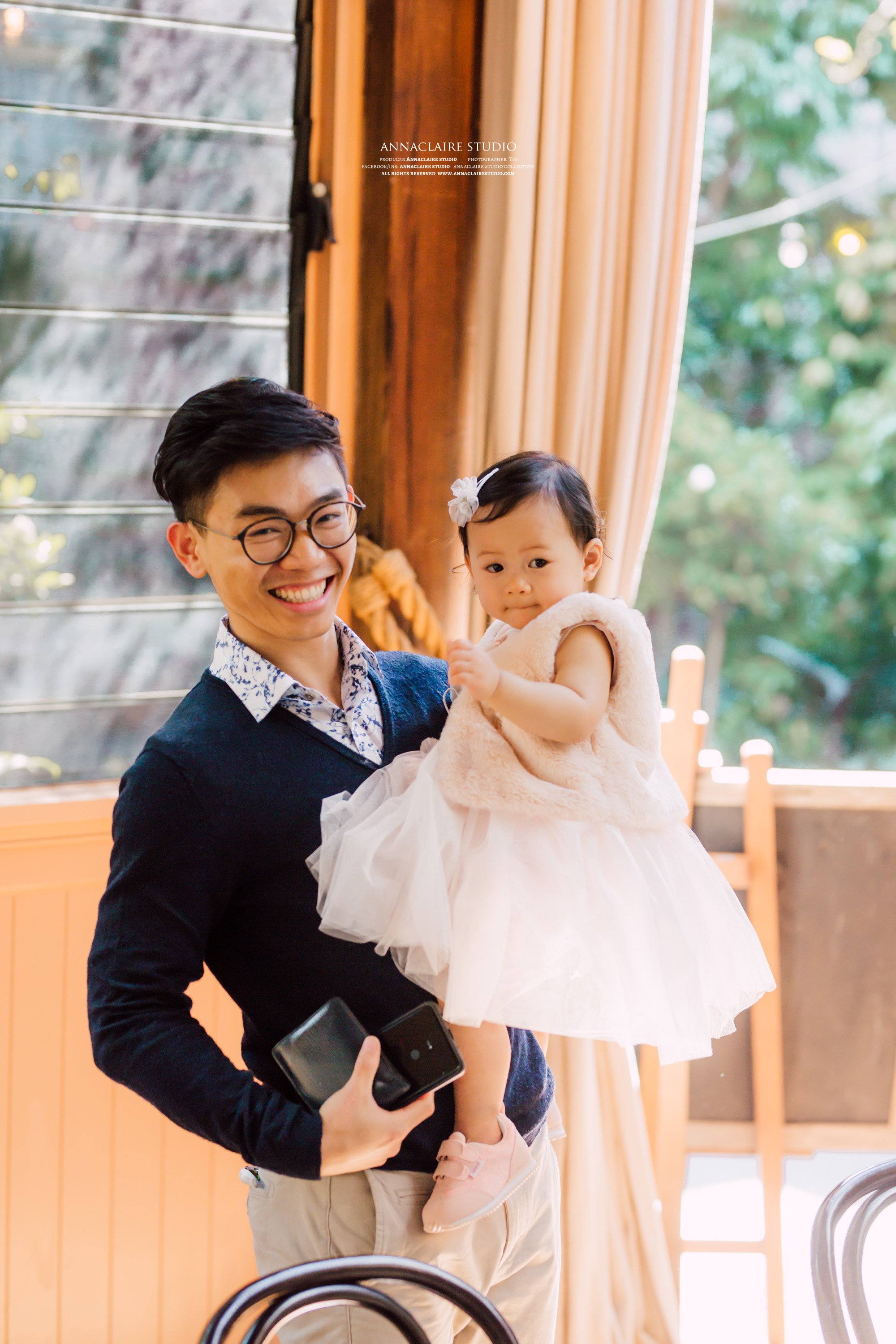 A man with glasses smiling while holding a young girl wearing a dress and a sleeveless fur vest indoors with large windows and curtains, with greenery outside.