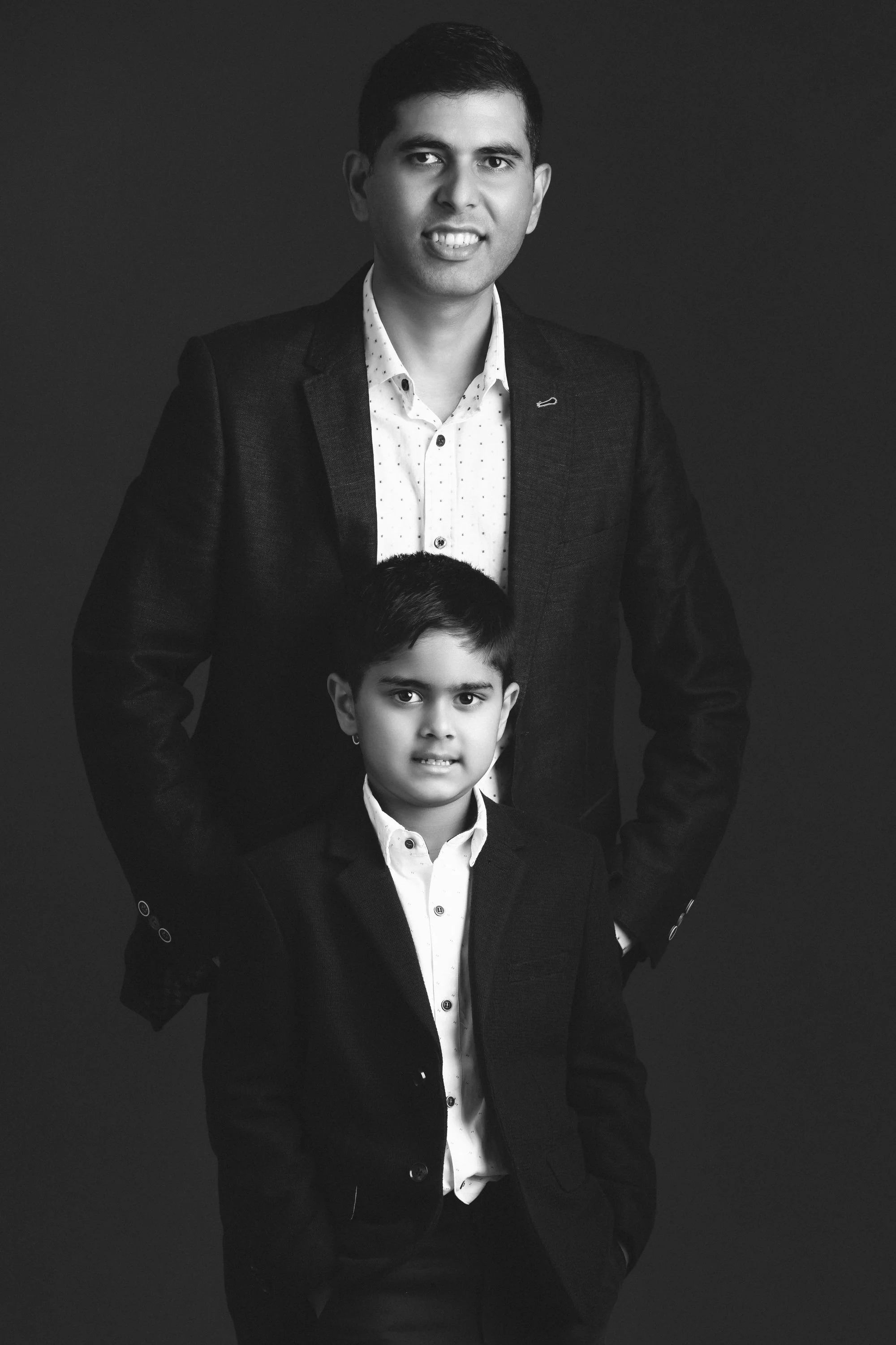 Black and white portrait of a man and a young boy, both wearing formal suits with white shirts, standing against a dark background.