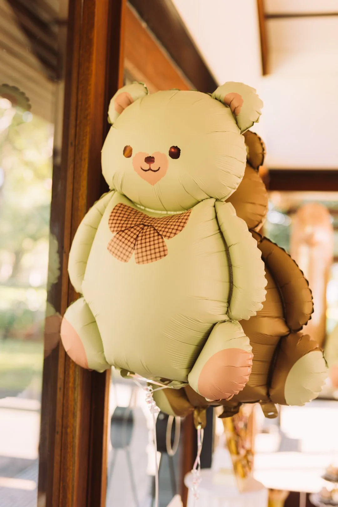 A light green balloon shaped like a teddy bear with a checkered bow tie, with a brown balloon panda behind it, hanging indoors near a wooden window.