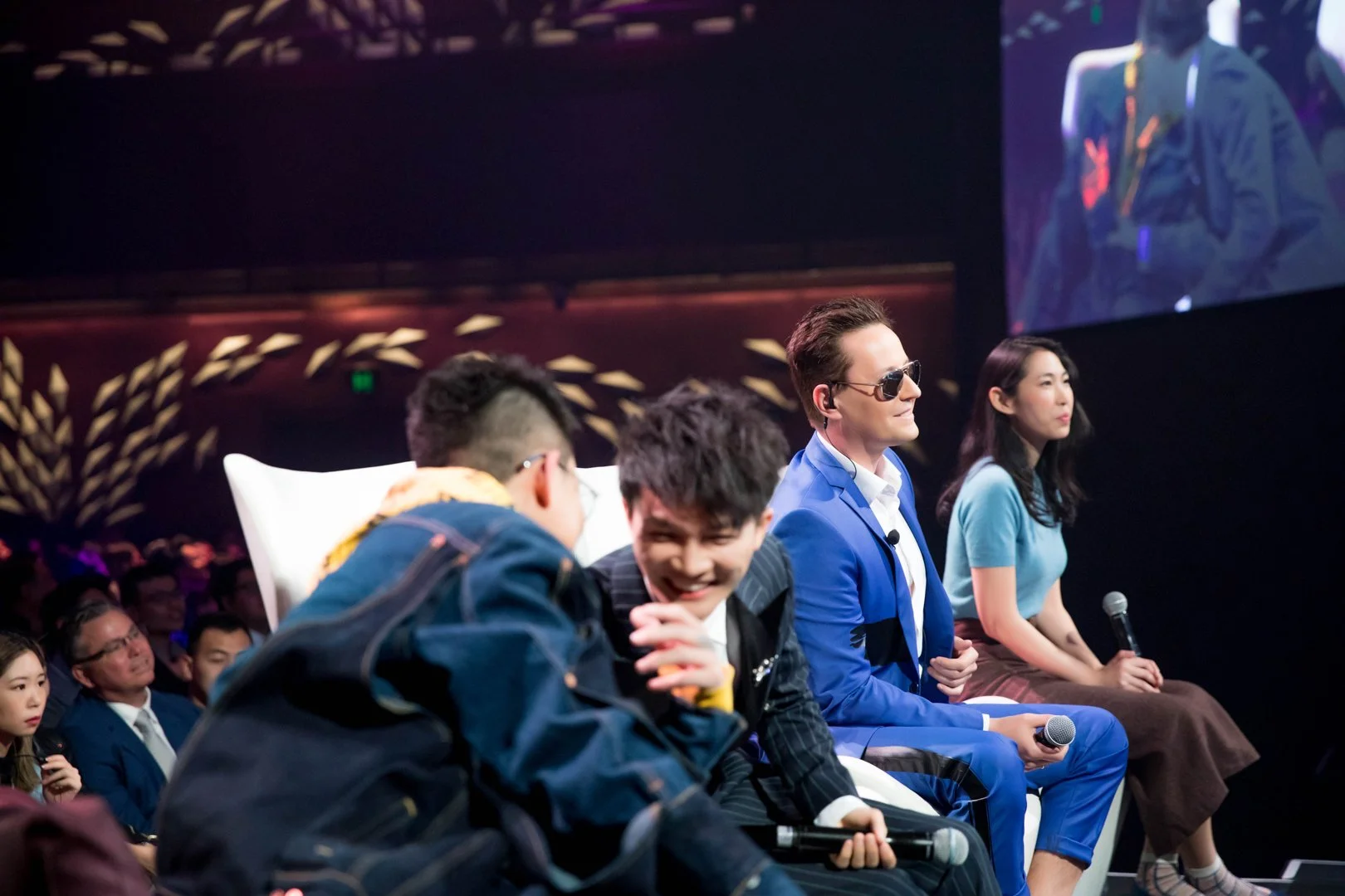 Panel of four people sitting on stage, with an audience in the background. The person second from the right is wearing sunglasses and a blue suit, holding a microphone. The other three individuals include a woman in a blue top and brown skirt, and tw