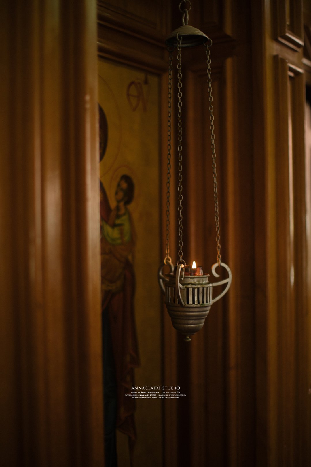 A hanging oil lamp with a lit flame, set against a wooden-paneled wall and a religious icon in the background.