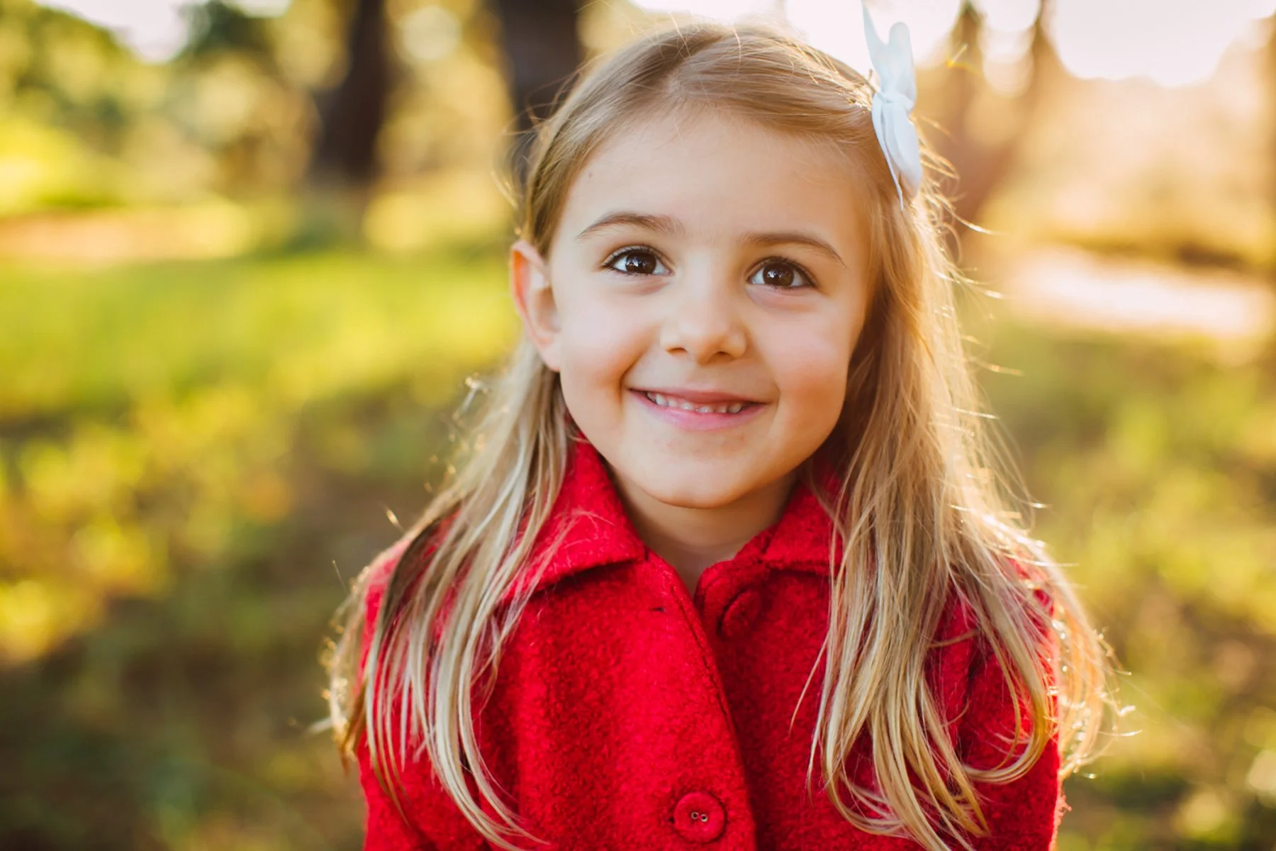A young girl with long blonde hair, wearing a red coat and a white bow in her hair, smiling outdoors in a park with sunlight filtering through trees.