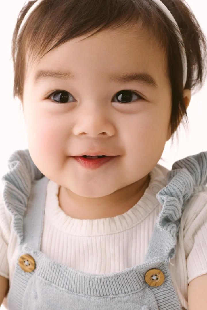 A young child with short brown hair, wearing a white headband and a light-colored outfit, smiling at the camera.