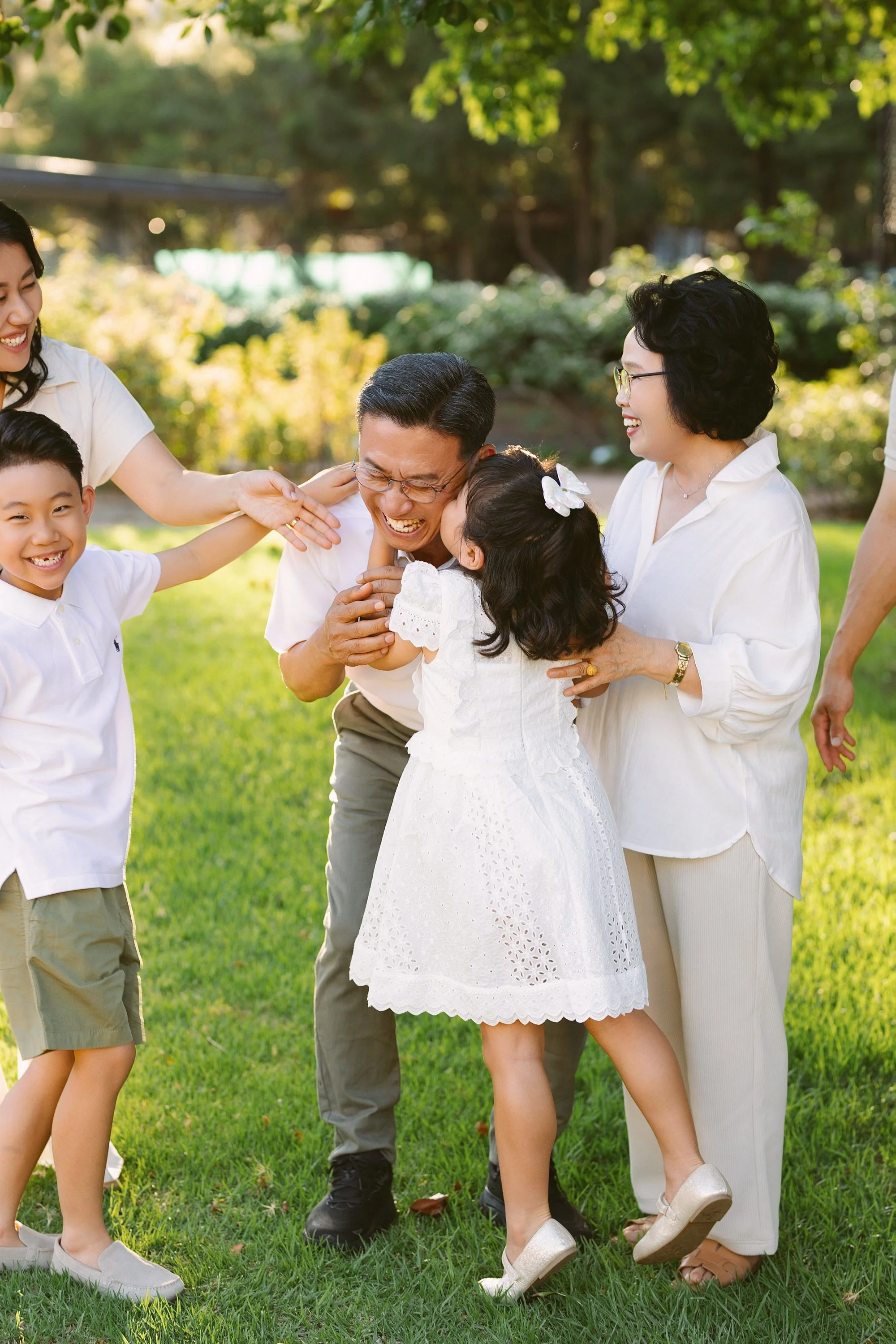 Family in a park laughing and hugging each other in bright sunlight, with green trees in the background. Adults and children are smiling and enjoying a joyful moment.