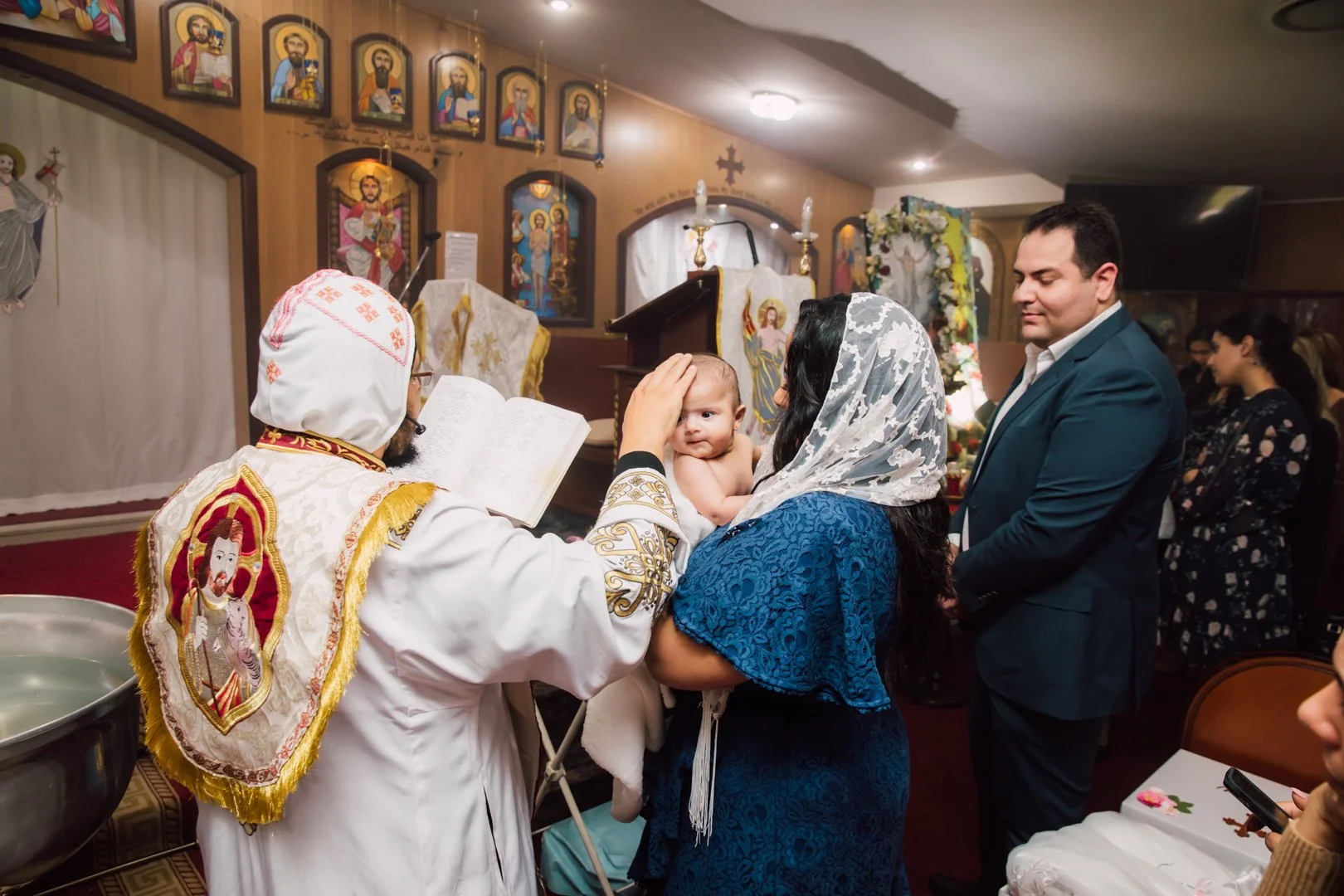 A mother holding her baby during a baptism ceremony in a church. The priest, dressed in white with religious symbols, touches the baby's forehead. The mother wears a white lace head covering and a blue dress. Next to her, a man in a suit and other fa