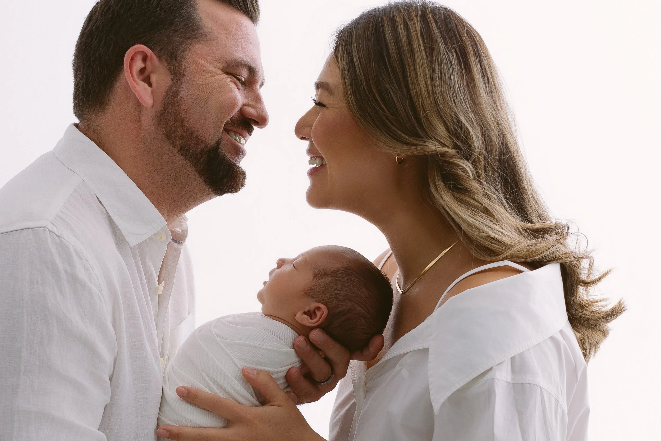 A smiling man and woman, holding a sleeping newborn baby between them, close to each other in a warm family moment.