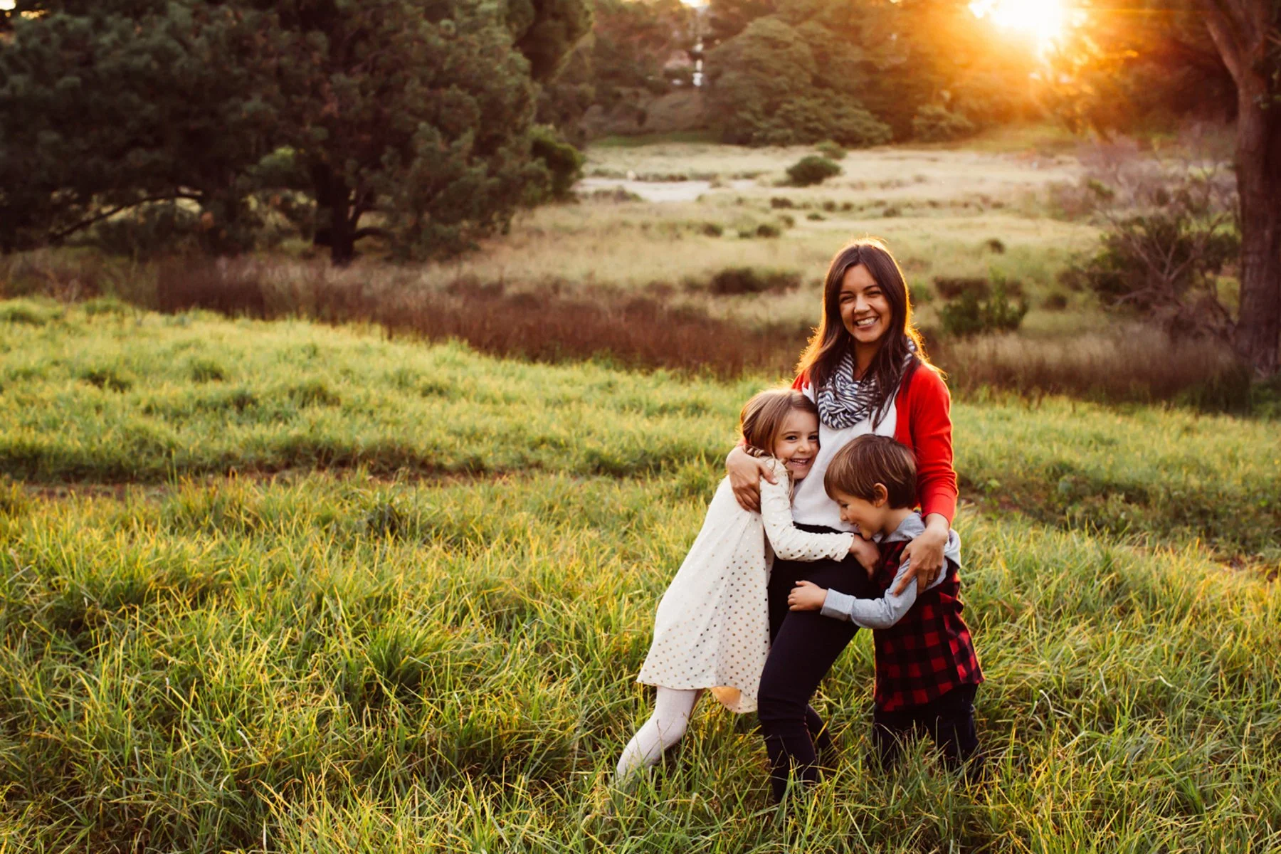 A mum with two kids standing outdoors in a grassy field at sunset, smiling and hugging each other.
