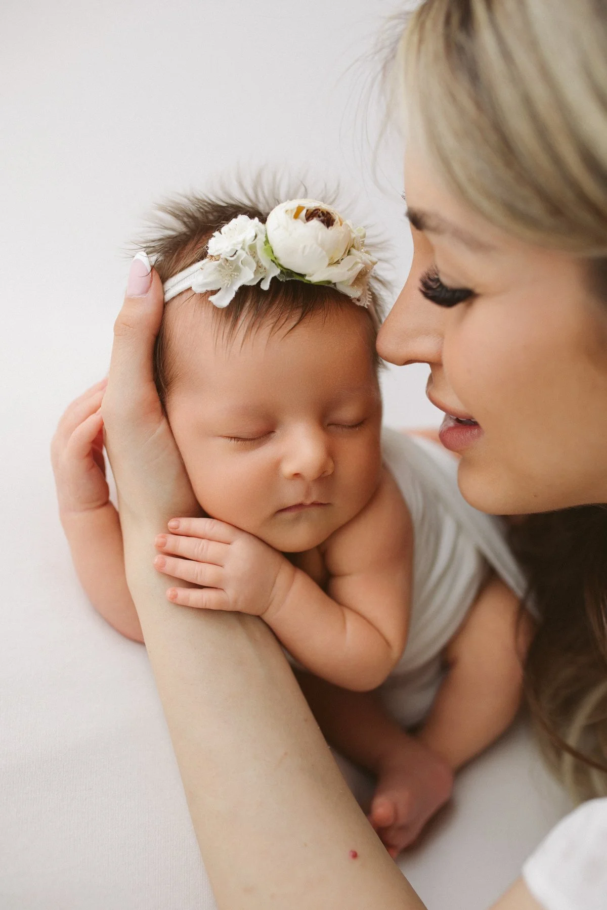 A woman gently cradles a sleeping newborn baby, who has a floral headband with white flowers and a small bee, close to her face in a tender moment.
