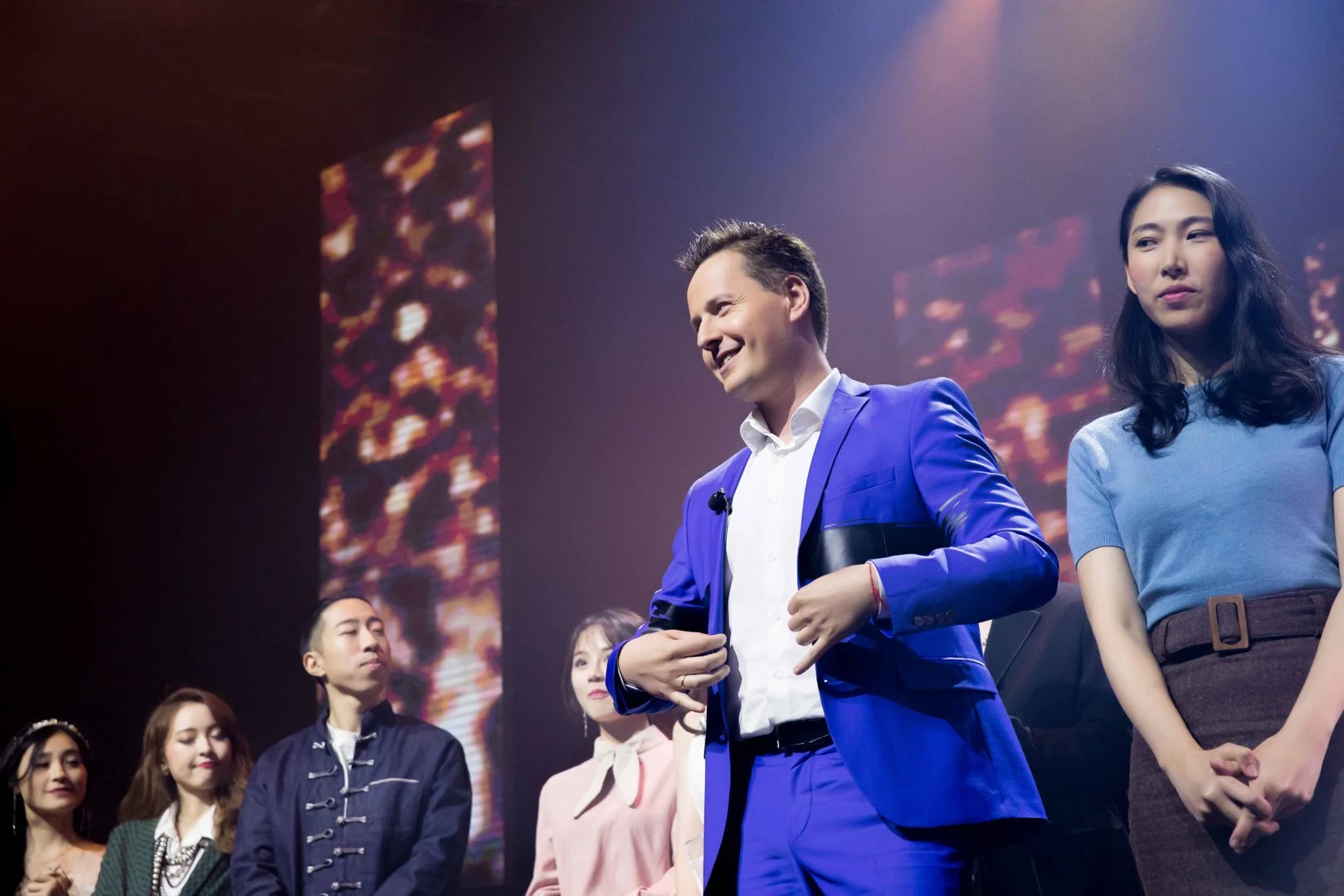 A group of people standing on a stage, with a man in a blue suit smiling and adjusting his jacket, several women and a man standing nearby, and colorful lights or visuals in the background.