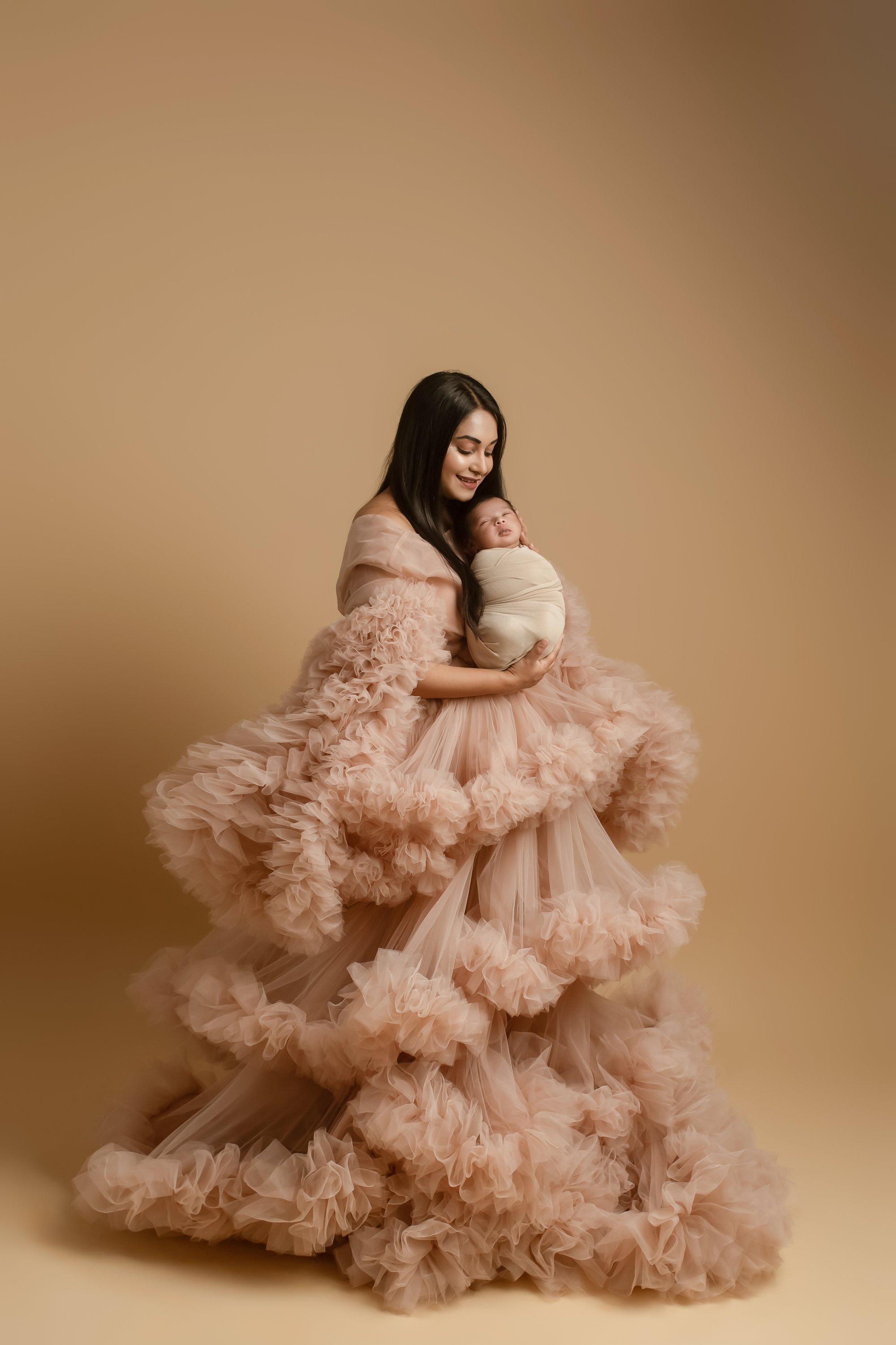 A pregnant lady in a voluminous, ruffled peach-colored gown holding a swaddled newborn baby against a beige background.