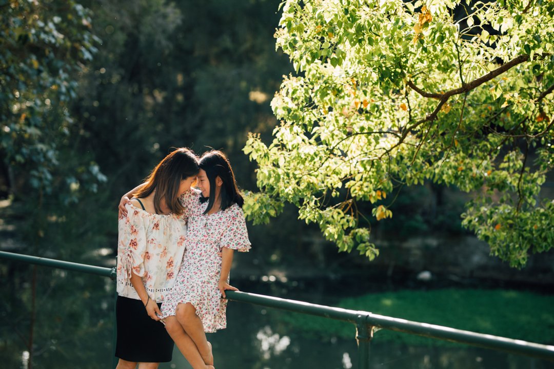 Two women sharing a close moment outdoors, leaning on a railing by a pond with green trees in the background.