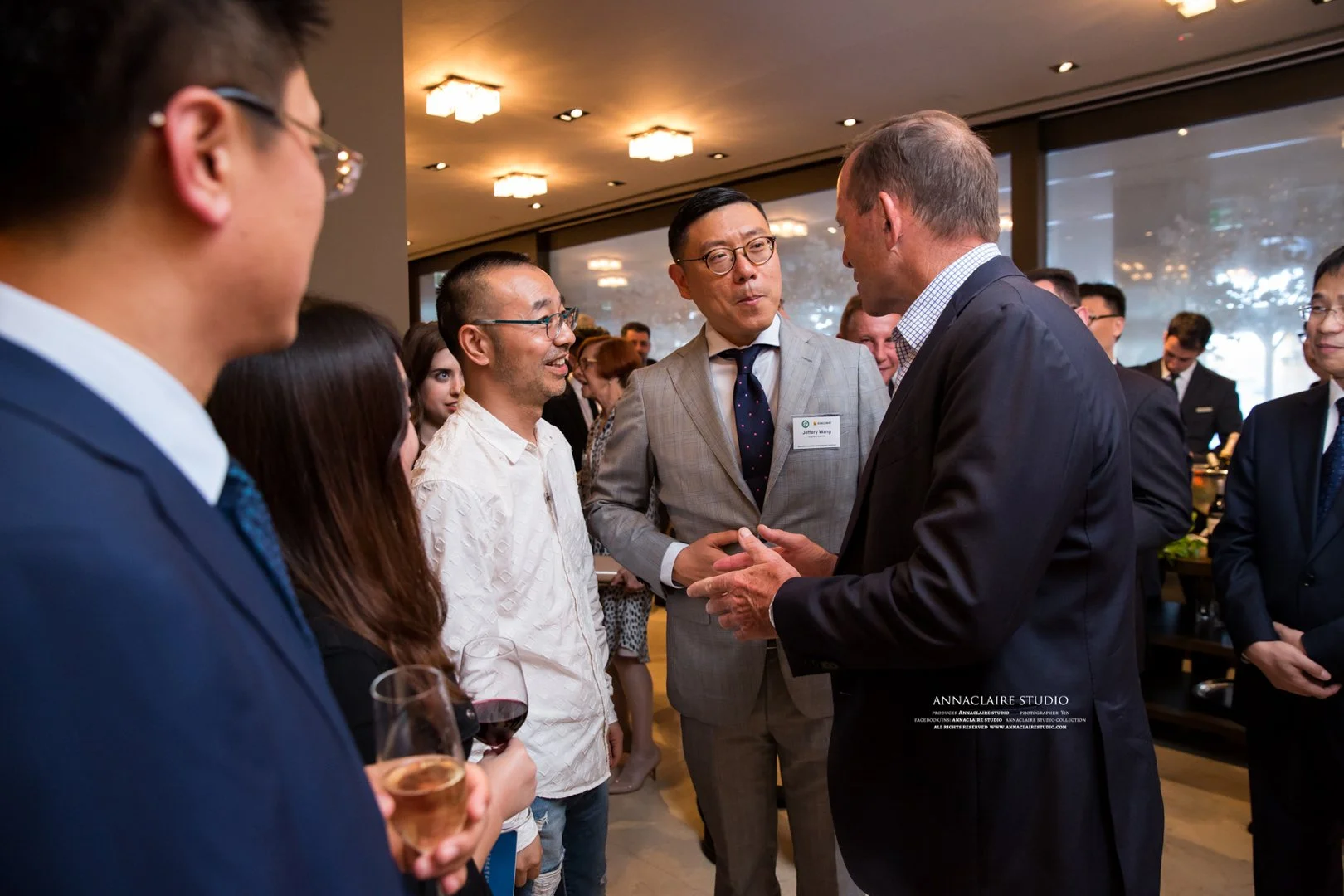 Group of business professionals engaged in conversation at a networking event, some holding glasses of wine, in an indoor setting with large windows and warm lighting.