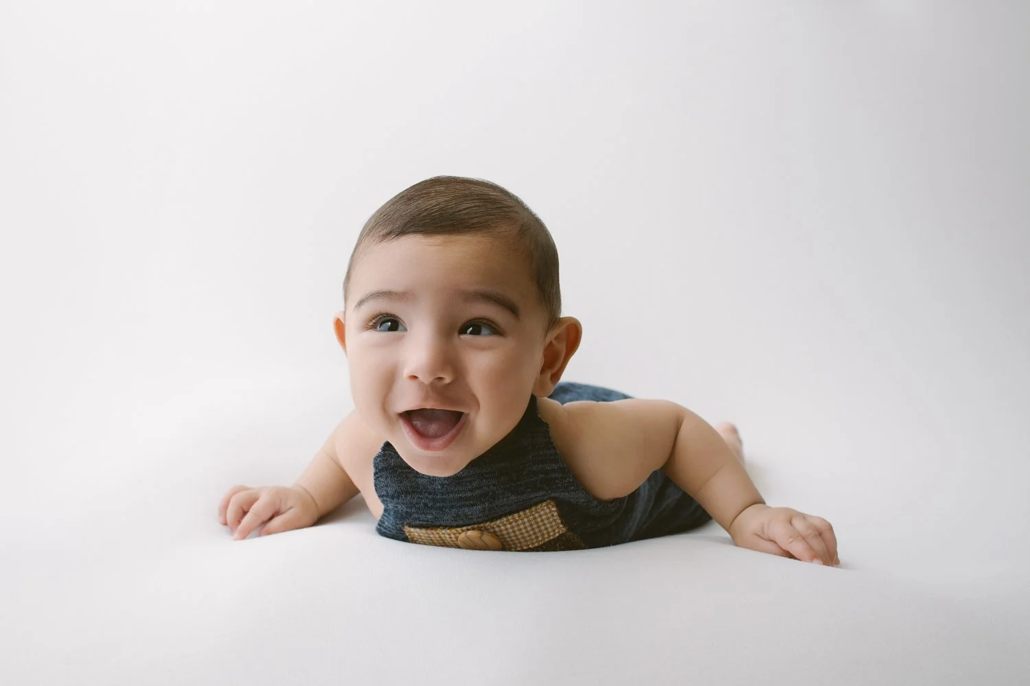 A happy baby lying on a white surface, smiling with open mouth, wearing a dark tank top.