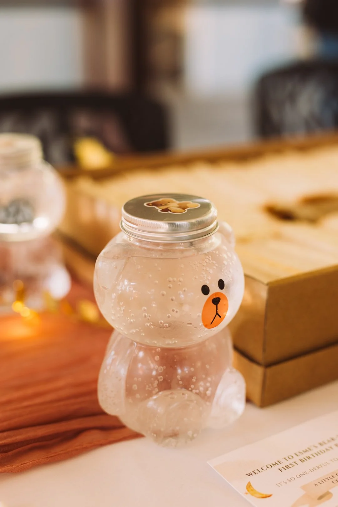 A bear-shaped clear plastic container with a face, filled with a bubbly light-colored beverage, sitting on a table near a wooden box.