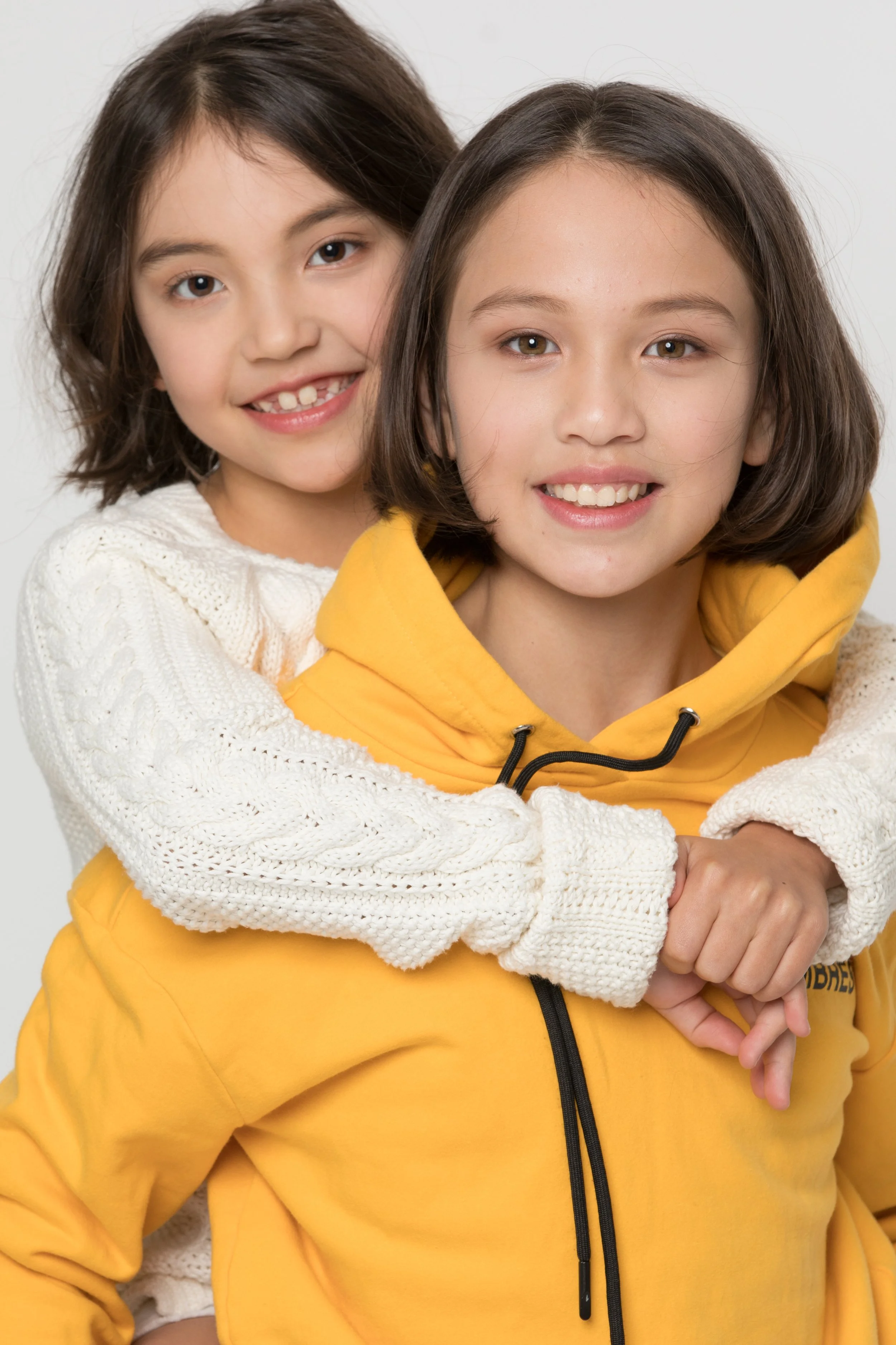Two young girls smiling, one standing behind the other with her arms wrapped around her shoulders. The girl in front is wearing a yellow hoodie, and the girl behind is wearing a white sweater. They look happy and close to each other.