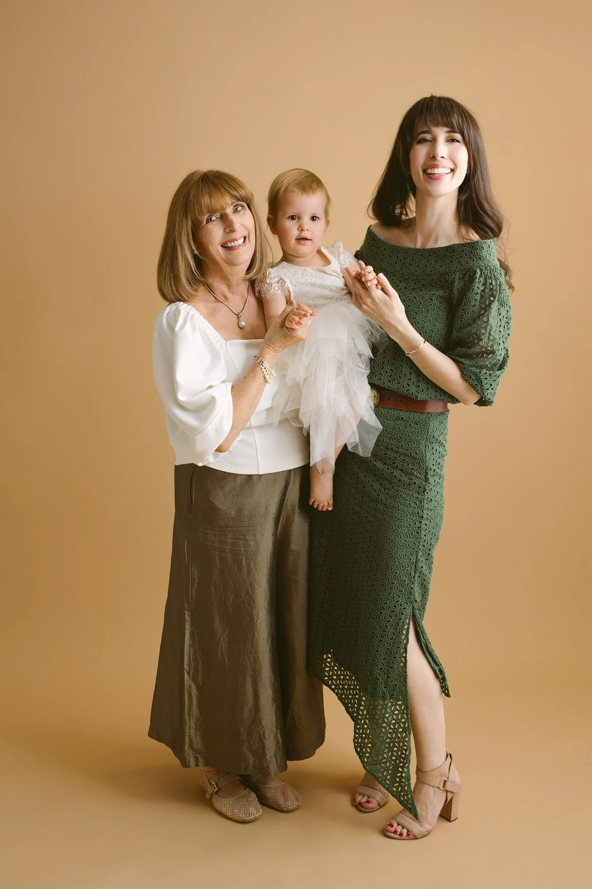 A woman with dark hair in a green dress holding a young girl in a white dress, standing next to an older woman with light hair in a white top and brown skirt, taken against a plain beige background.