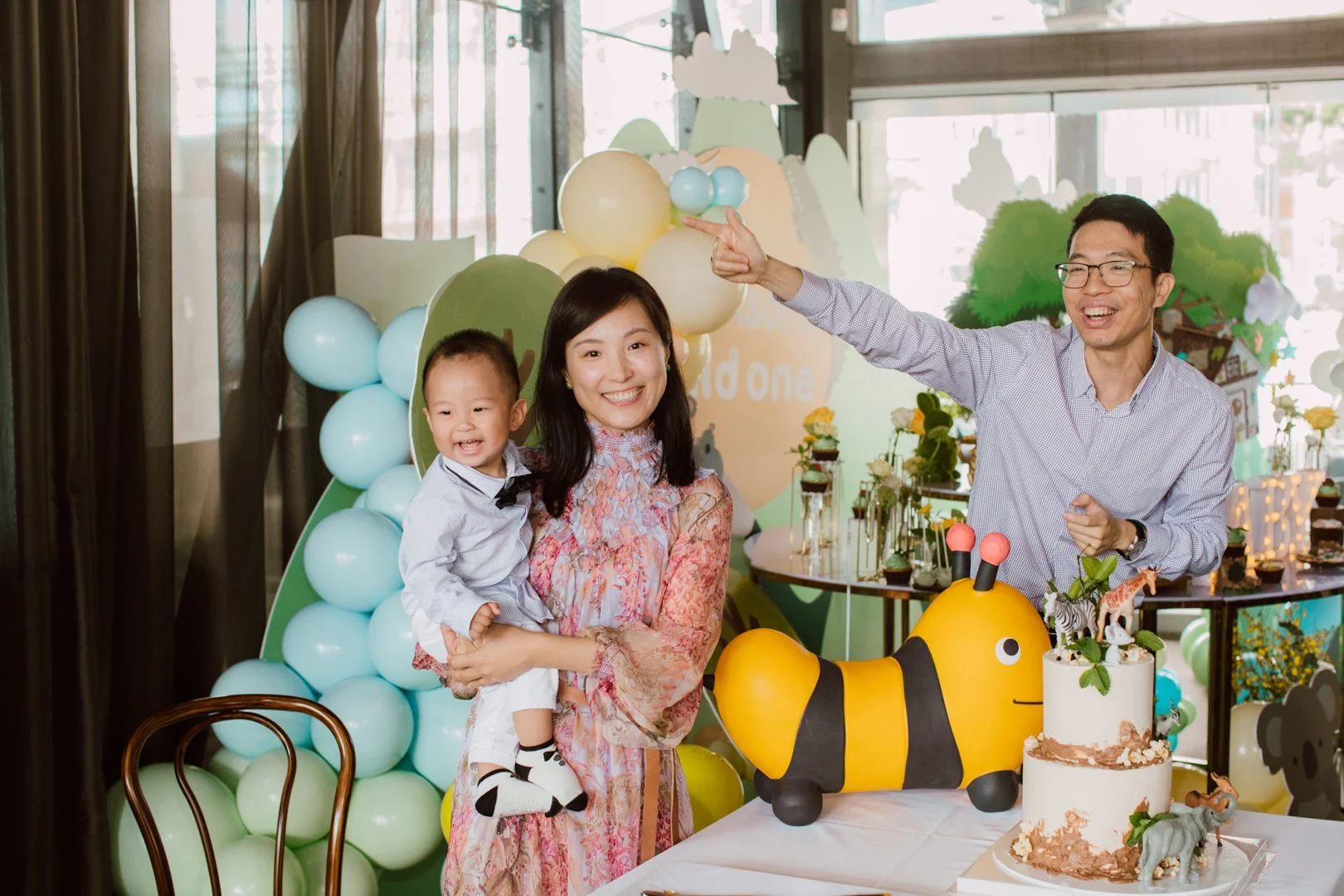 Family celebrating a child's first birthday with decorations including balloons, a cake with animal figurines, and a large bee-shaped decoration, in an indoor setting with large windows.