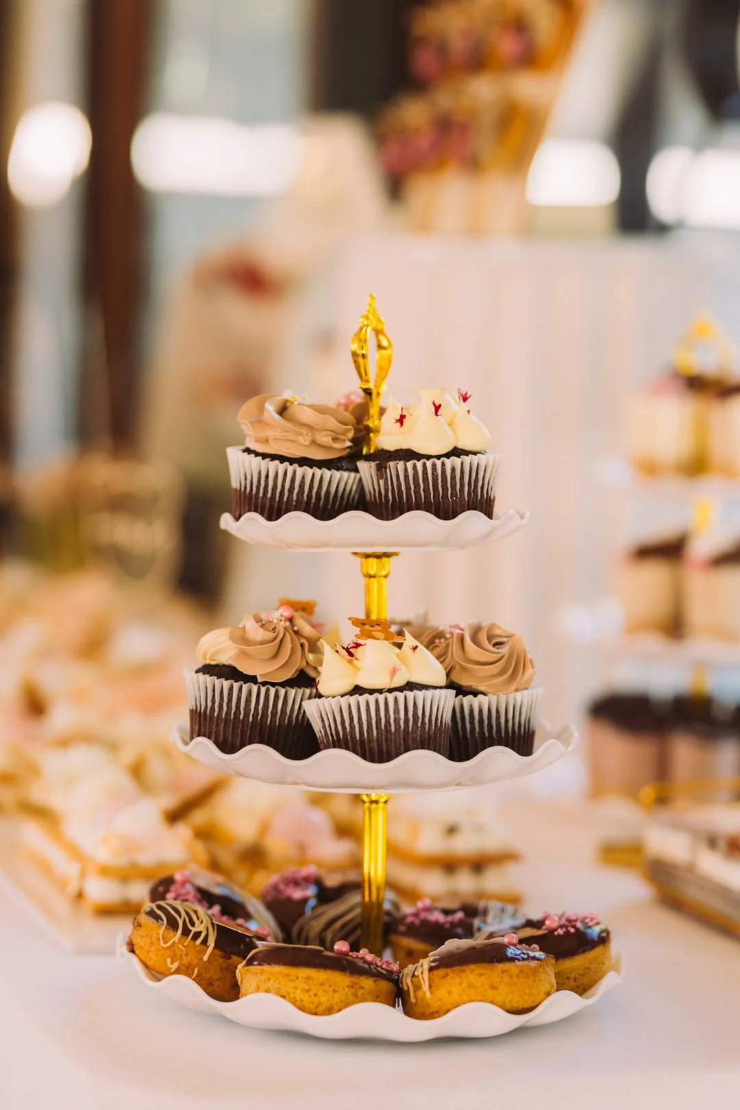 A three-tiered dessert stand with assorted cupcakes and cookies, decorated with chocolate and cream, displayed on a table at a celebration or event.