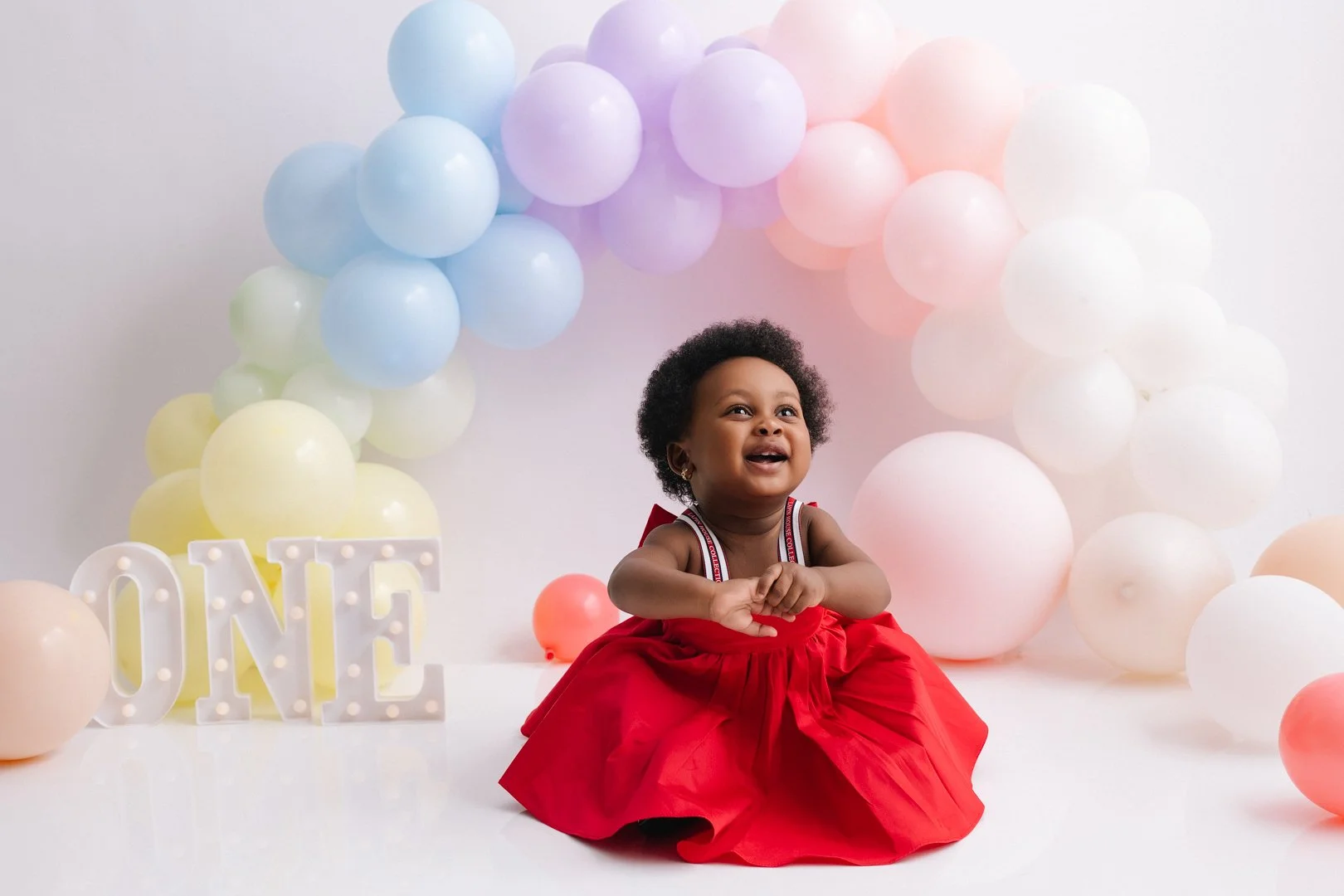A smiling 1 yr old  girl with curly hair in a red dress sitting on a white surface, surrounded by pastel-colored balloons in a semi-circle and white decorative letters spelling 'ONE' at a birthday celebration.