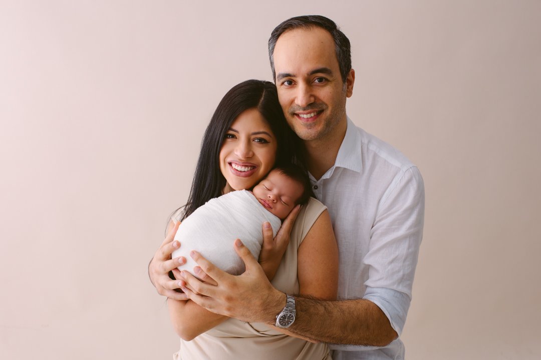Smiling young couple holding a sleeping newborn wrapped in a white blanket against a plain background.