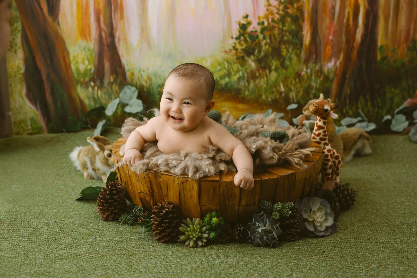 A happy baby sitting in a woodland-themed setting with plush animals, pinecones, and succulents, against a painted forest backdrop.