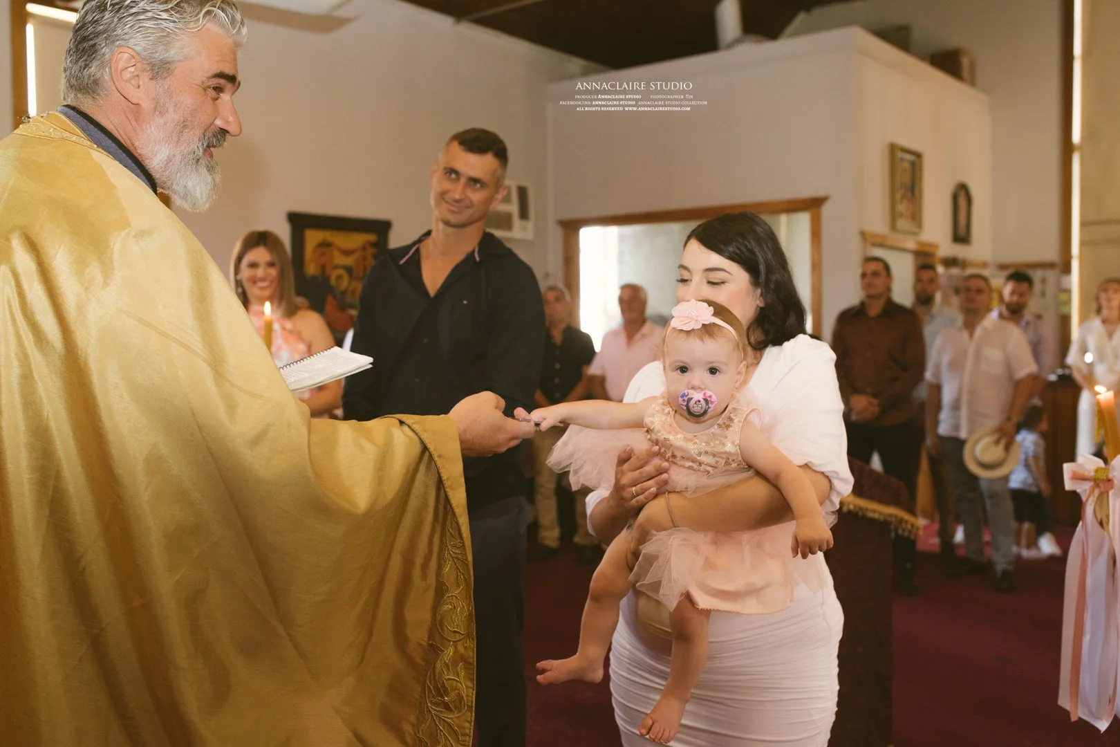 A baby girl with a pacifier in her mouth, dressed in a peach dress, is held by a woman during a baptism ceremony. The woman is handing the baby to a priest dressed in a gold robe. Several people are observing the event in the background.