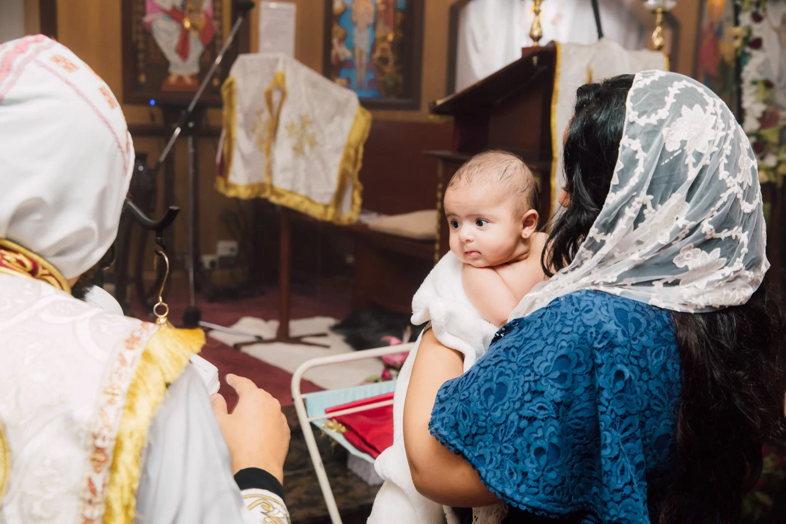 A woman holding a baby during a religious ceremony with an officiant in traditional vestments present.