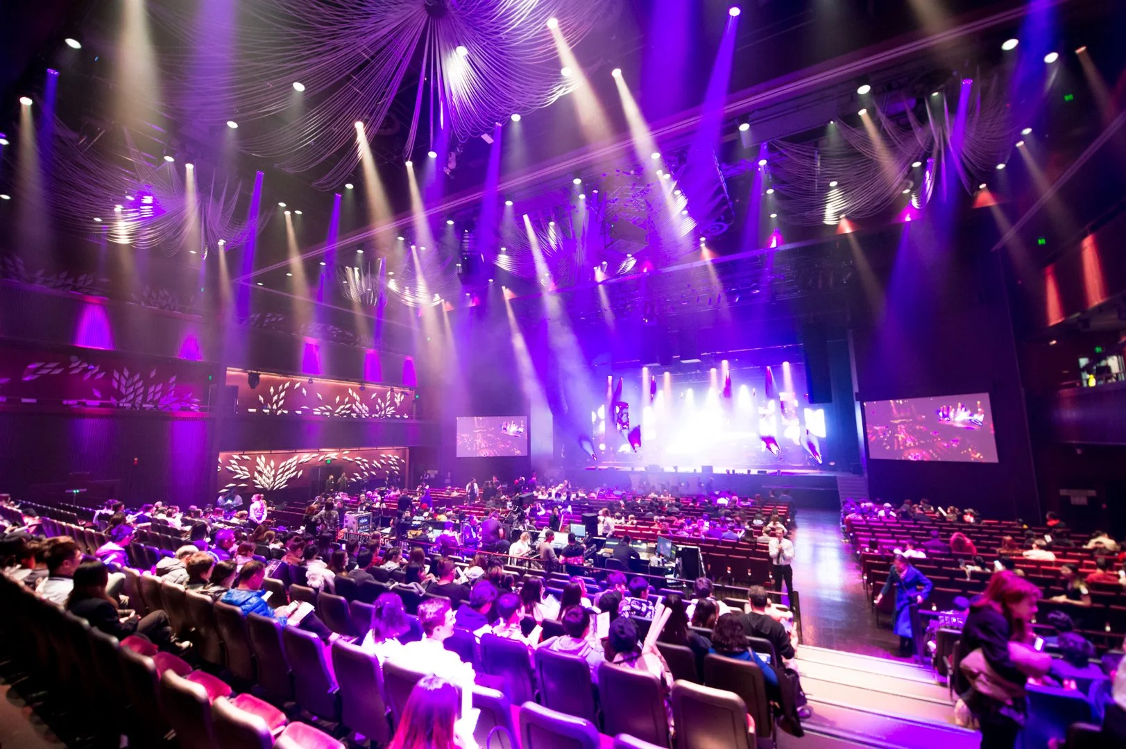 An indoor auditorium with an audience seated in front of a stage. The stage is brightly lit with purple and white lights, and two large screens display visuals. The ceiling is decorated with hanging fabric and dynamic lighting effects, creating a vib