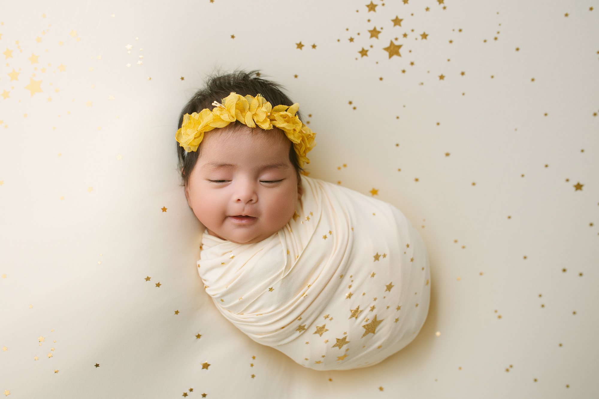 A newborn baby wearing a yellow floral headband, wrapped in a cream blanket with gold star confetti, resting on a cream surface with gold star confetti scattered around.