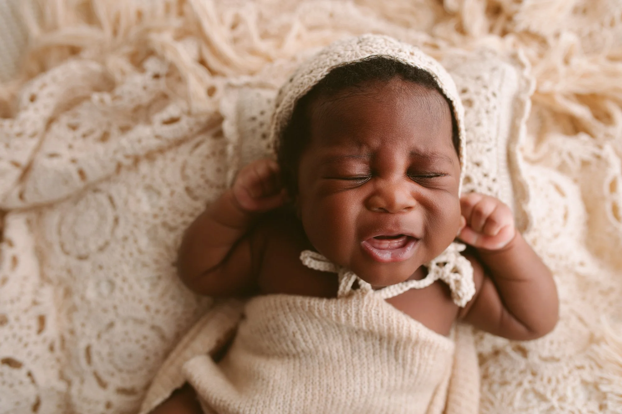 Close-up of a crying baby , wearing a knit bonnet and wrapped in a cream-colored blanket, lying on a boho textured cream-colored crochet blanket.