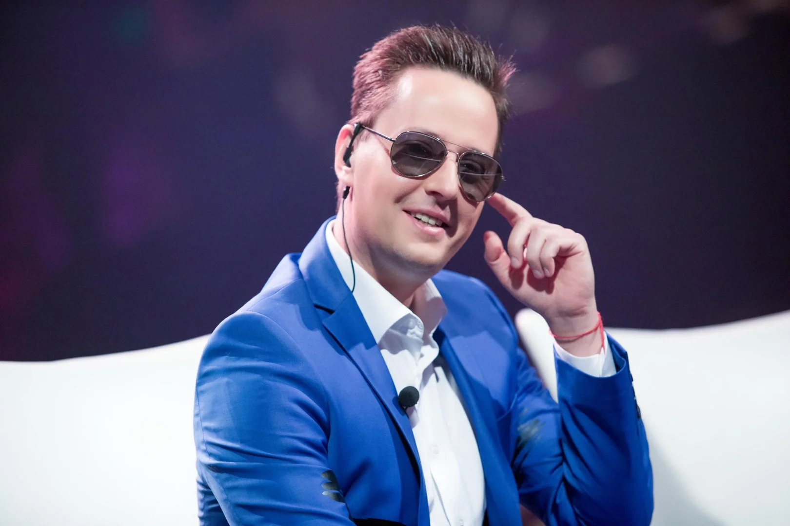 Man with brown hair wearing sunglasses and a blue suit, smiling and listening through an earpiece, sitting on a white chair.