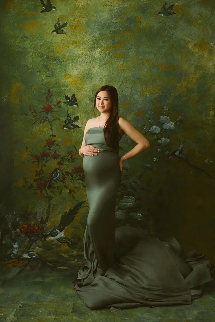 A woman in a long, elegant green dress standing in front of a painted backdrop of birds and trees.