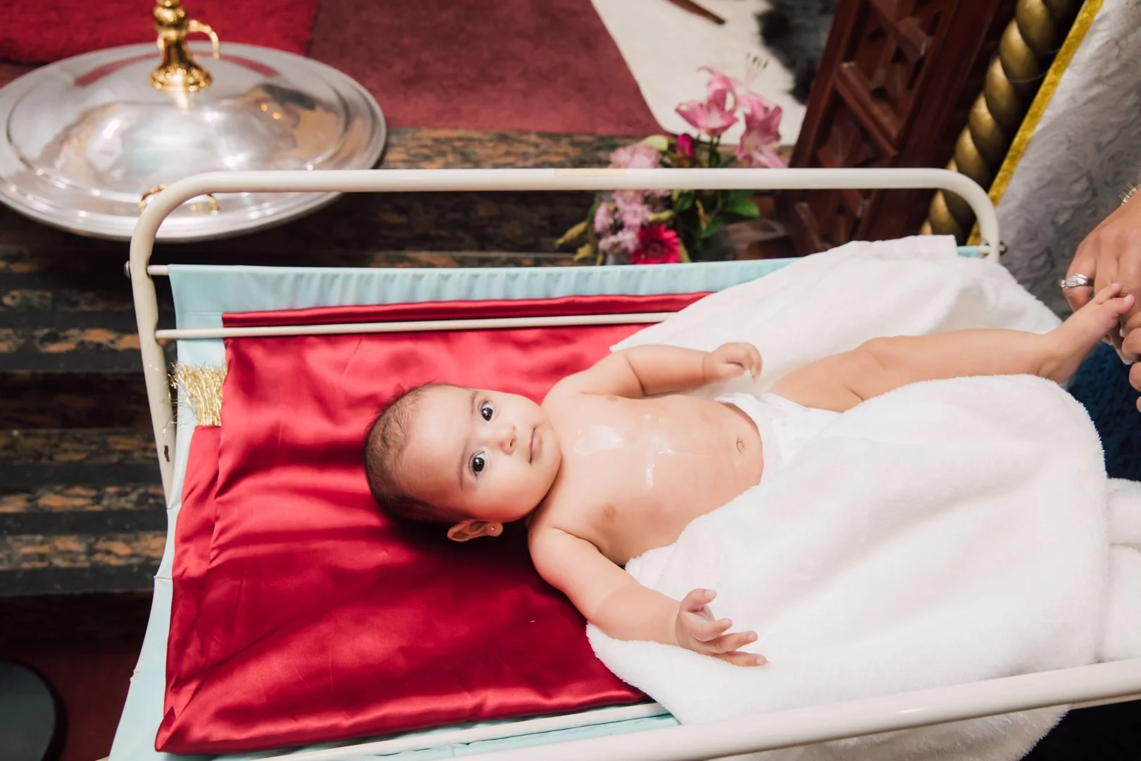 A baby lying in a hospital bed with a white blanket, looking at the camera, with a patch on its chest, holding an adult's hand.