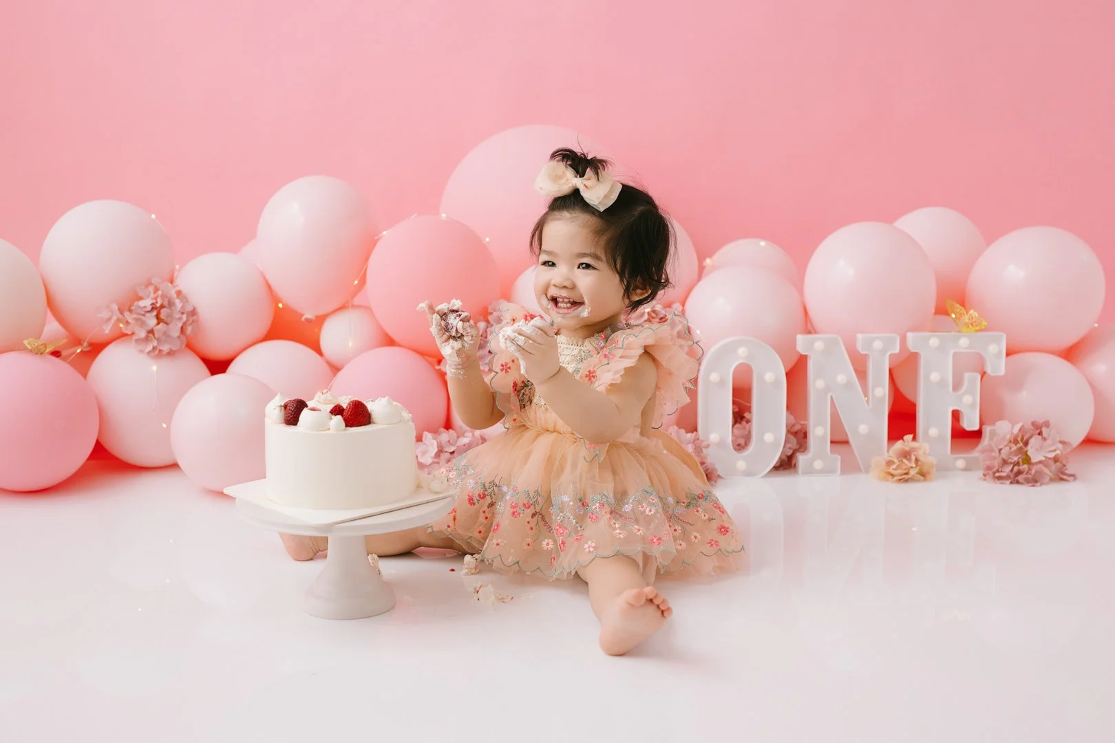 A  one year old babysitting on the floor celebrating her first birthday with a cake, surrounded by pink balloons, flowers, and illuminated letter signs spelling 'ONE' against a pink background.