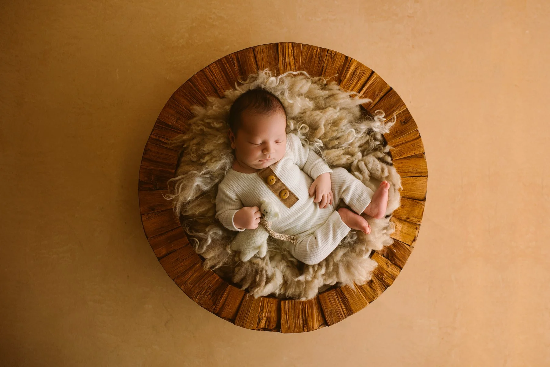 A sleeping baby lying on a circular wooden platform, surrounded by soft woolly material, holding a small stuffed animal.