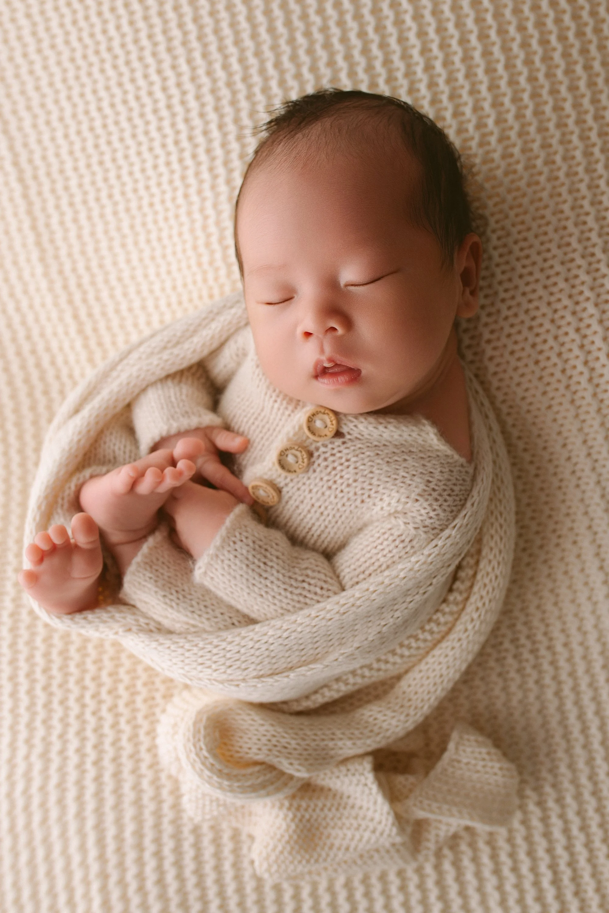 A sleeping infant wrapped in a cream-colored knitted blanket, lying on a matching textured blanket.
