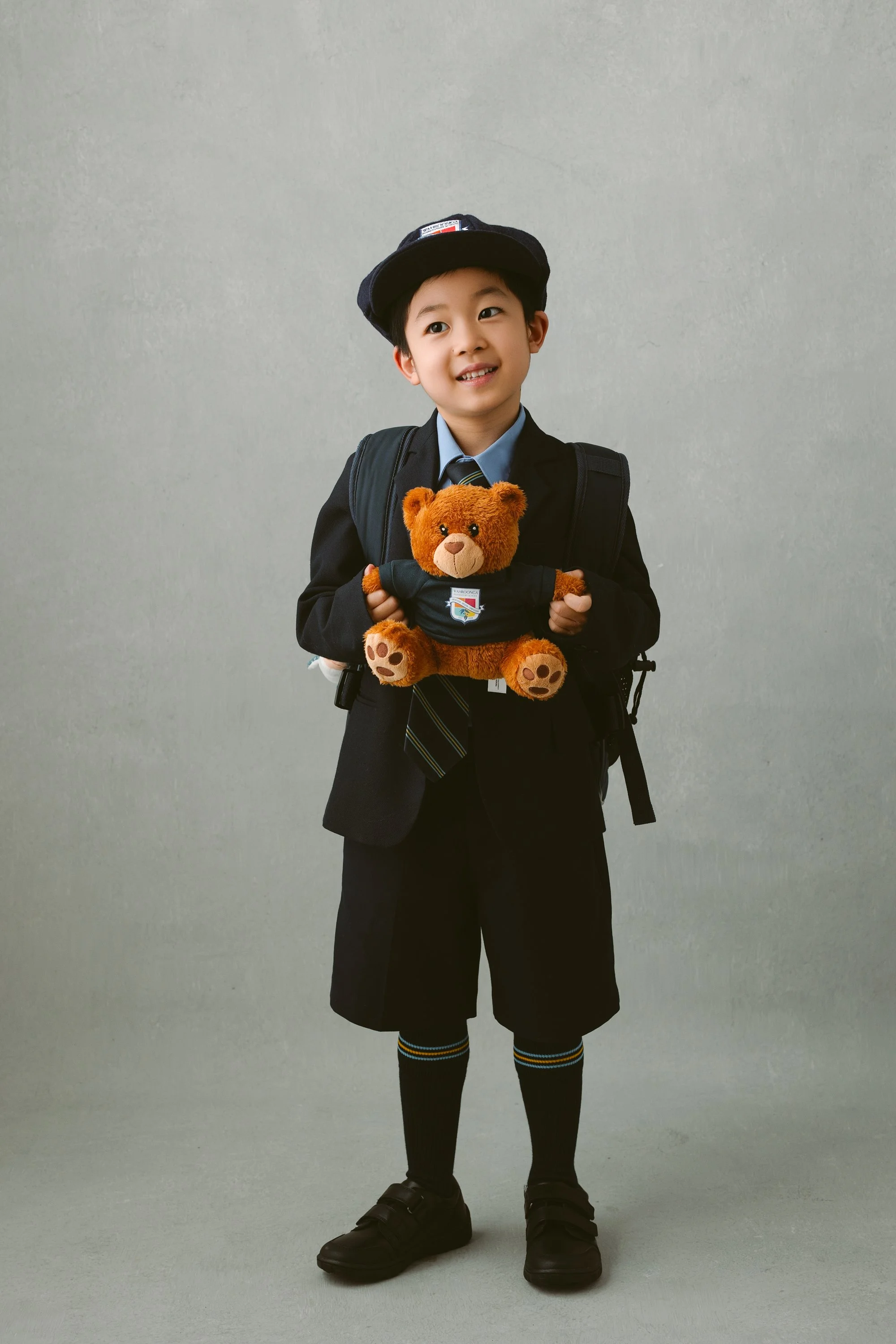 Young boy in school uniform holding a teddy bear, standing against a plain background.