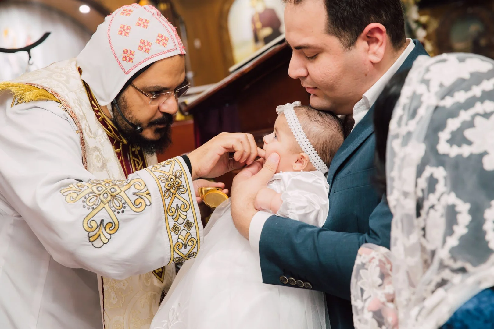 A priest in traditional attire performs a religious ceremony with a baby held by an adult man. The priest is touching the baby's mouth with his fingers during the ceremony. The adult man is holding the baby close to his chest, both are looking attent
