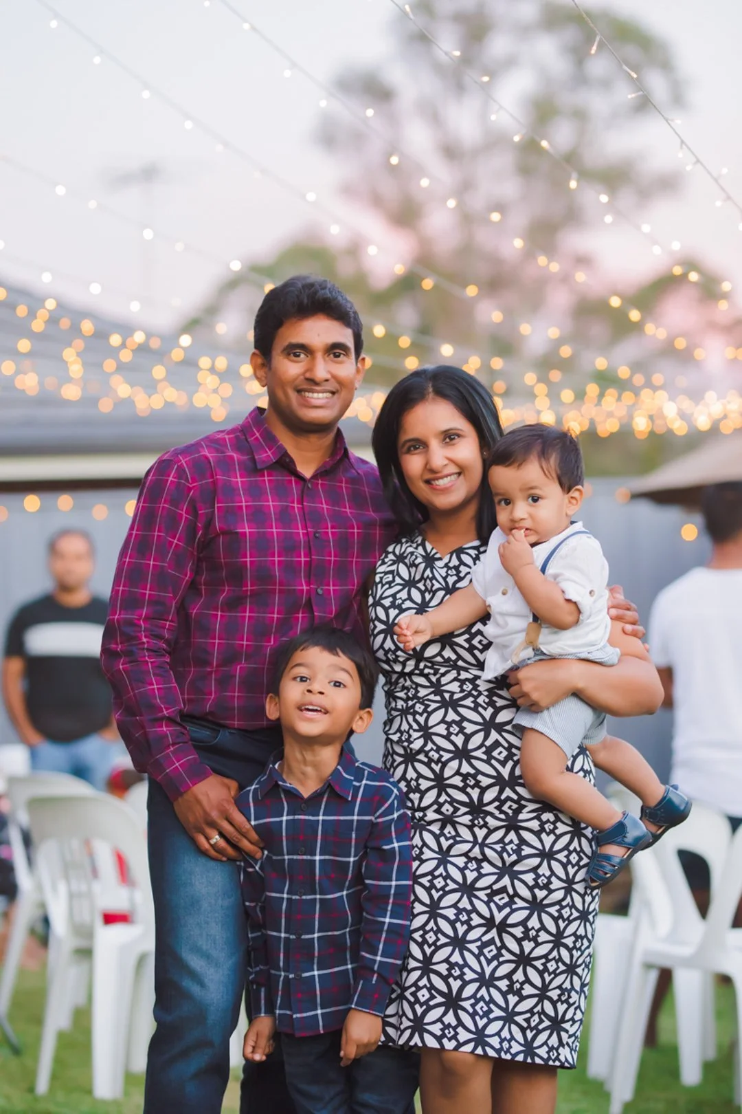 A happy family of four, two adults and two children, posing outdoors at a celebration with string lights in the background, smiling at the camera.