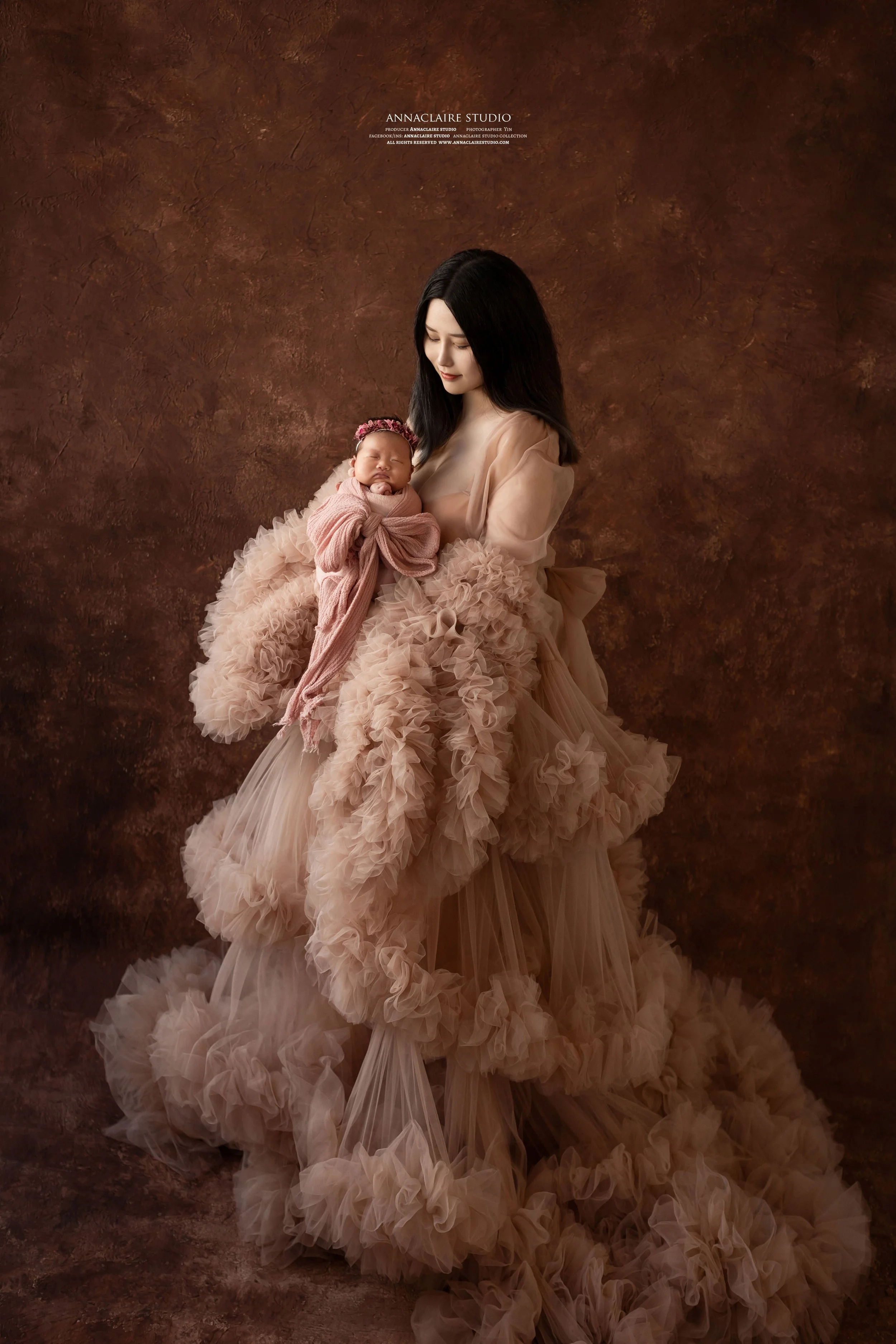 A new mum holding a sleeping baby against a textured brown background. The woman wears a flowing, ruffled, beige gown, and the baby is dressed in pink with a matching headband.