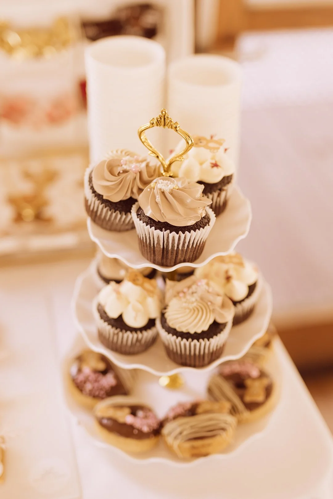 A tiered display of assorted decorated cupcakes with chocolate and vanilla frosting, topped with decorative gold and pink accents, on a white cake stand.