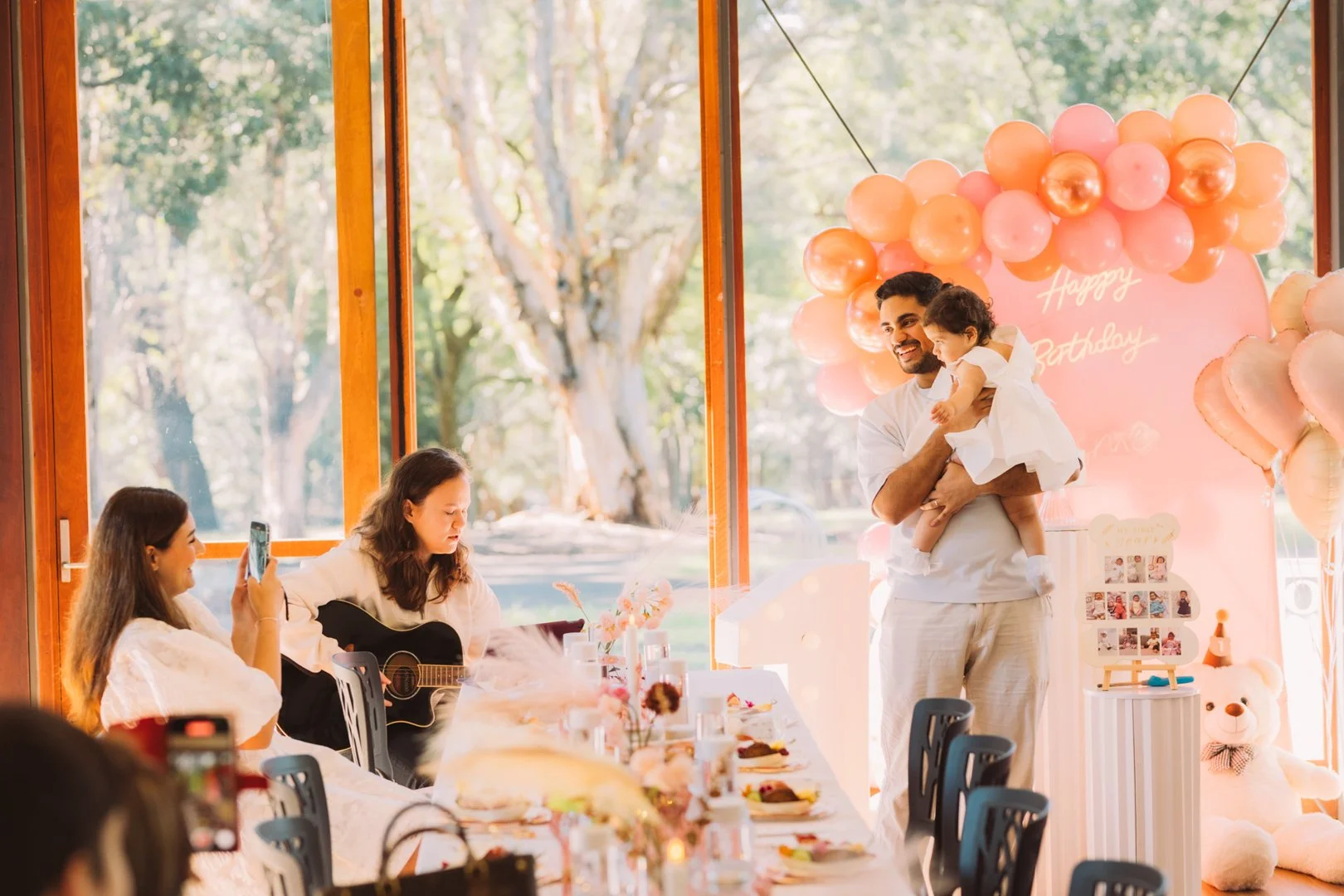 A family celebrating a child's birthday with pink and peach balloon decorations, a teddy bear, and a setting with food and drinks, with a man holding a young girl while another woman plays guitar and a girl recording with her phone.